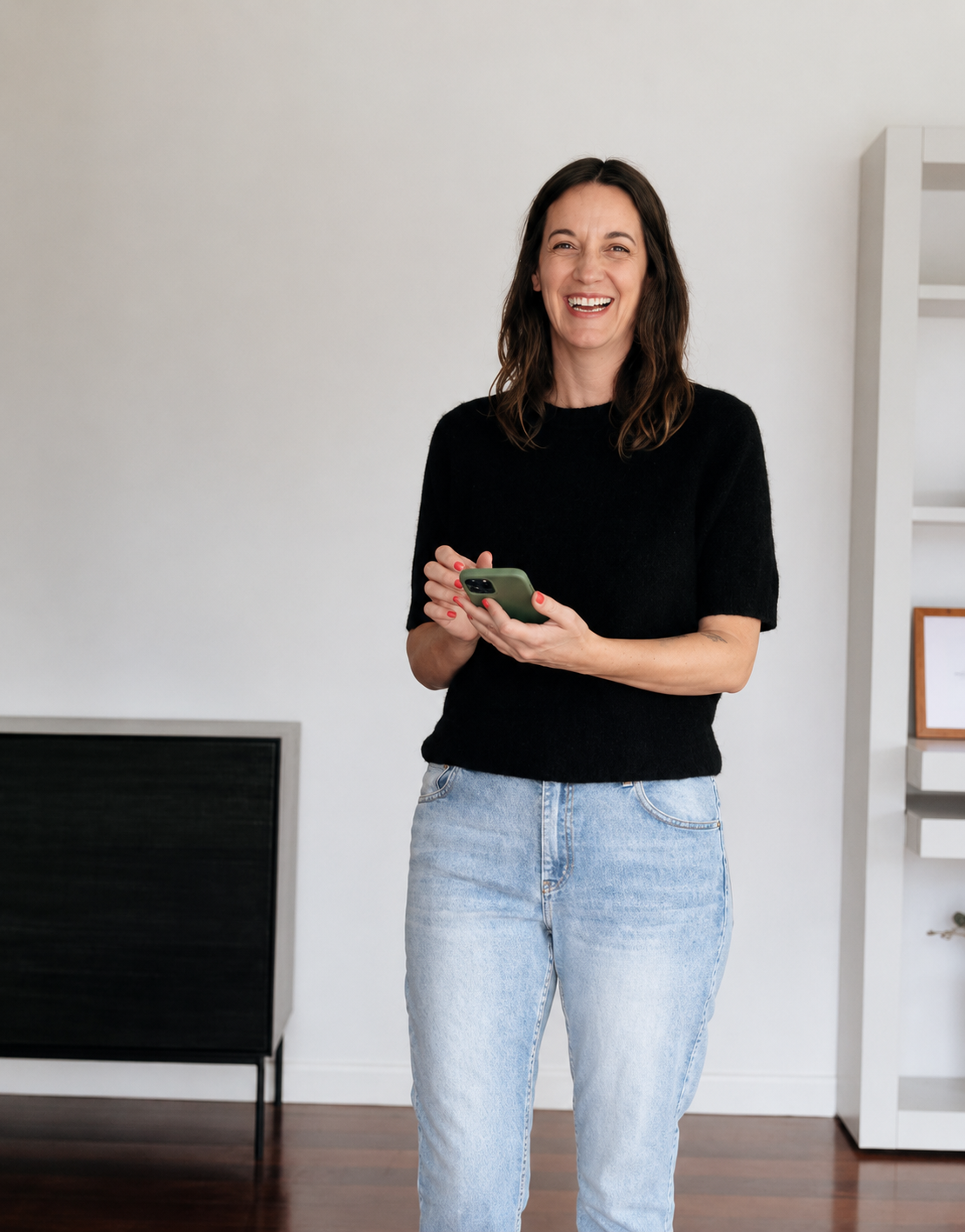 Emma from Aura studio smiling while holding a smartphone in a modern, minimalistic room with white walls and a dark wood floor.