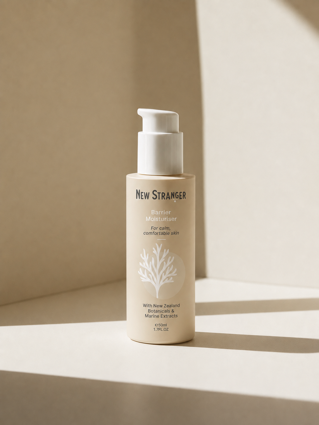 A beige bottle of New Stranger Barrier Moisturiser with a pump, placed on a light-colored surface with shadows and background created by natural light.