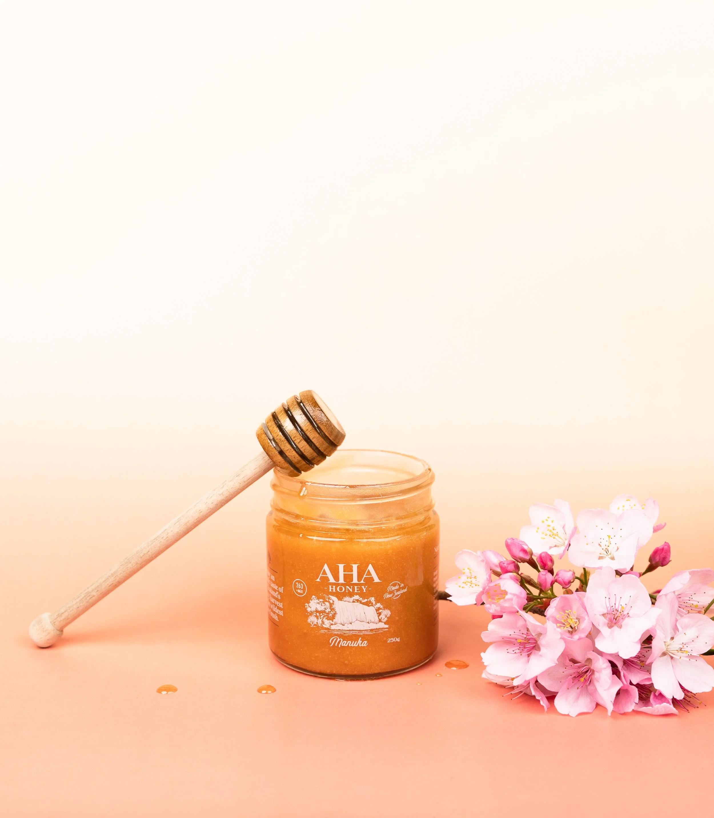 Product photography of an AHA Honey Manuka honey jar styled with a honey dipper and fresh flowers, showcasing premium New Zealand honey packaging.