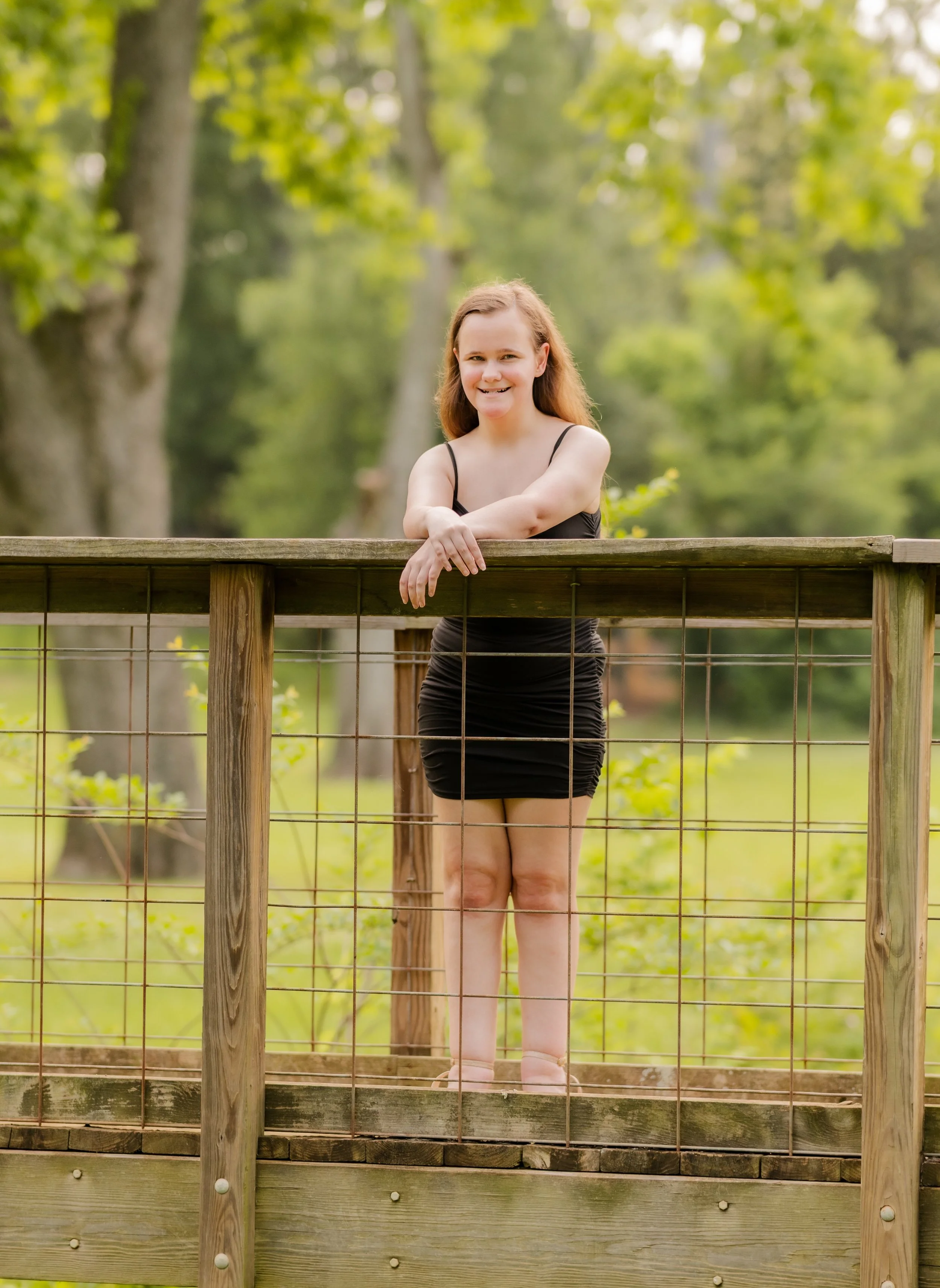 A woman in a black dress standing behind a wooden fence with wire mesh, in a park or wooded area with green trees in the background.