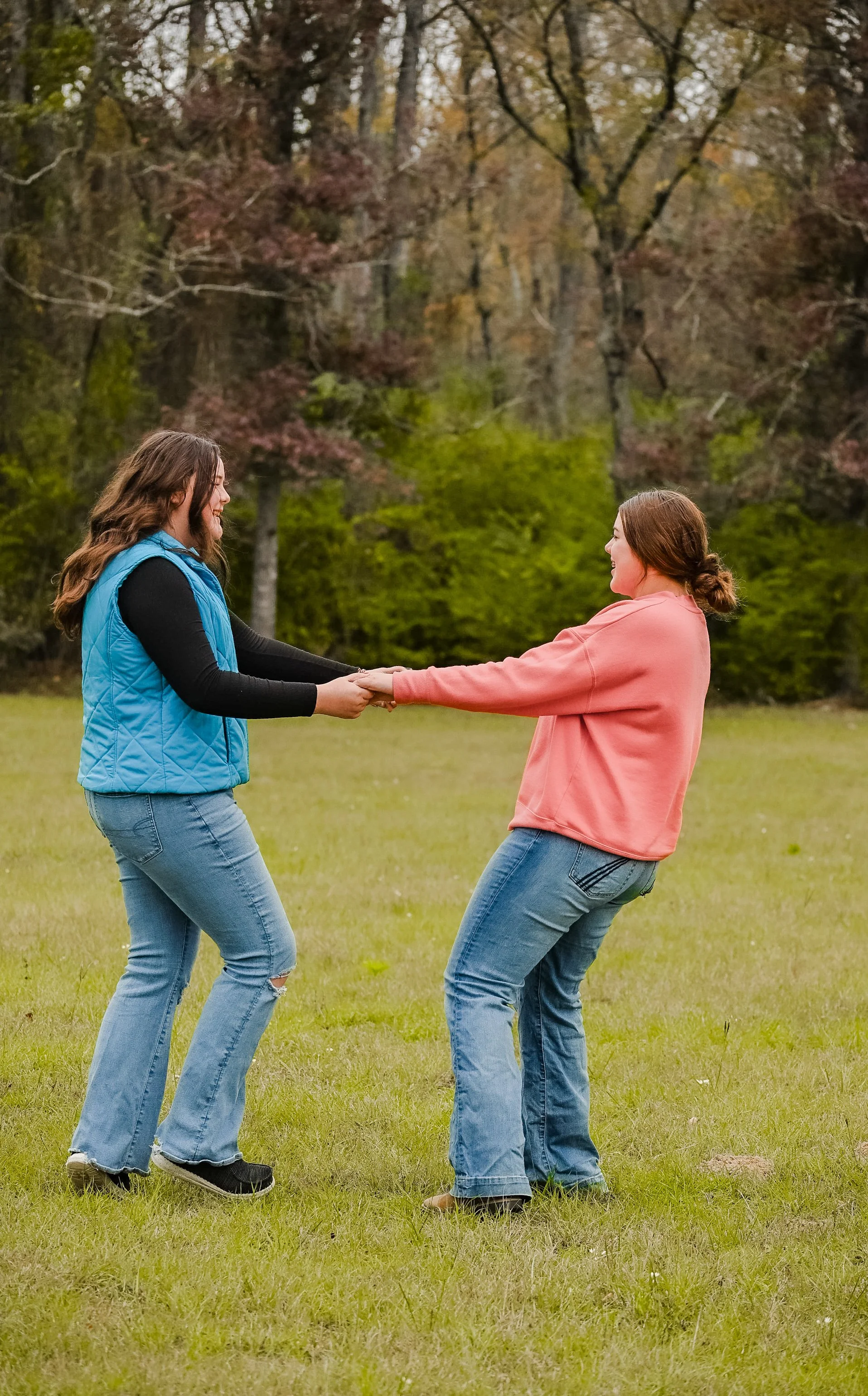 Two women are playing a game of tug of war outdoors in a grassy area, with trees and autumn foliage in the background. One woman has long brown hair, wears a blue vest over a black shirt, and blue jeans. The other woman has brown hair tied back, wears a pink sweatshirt, and blue jeans.