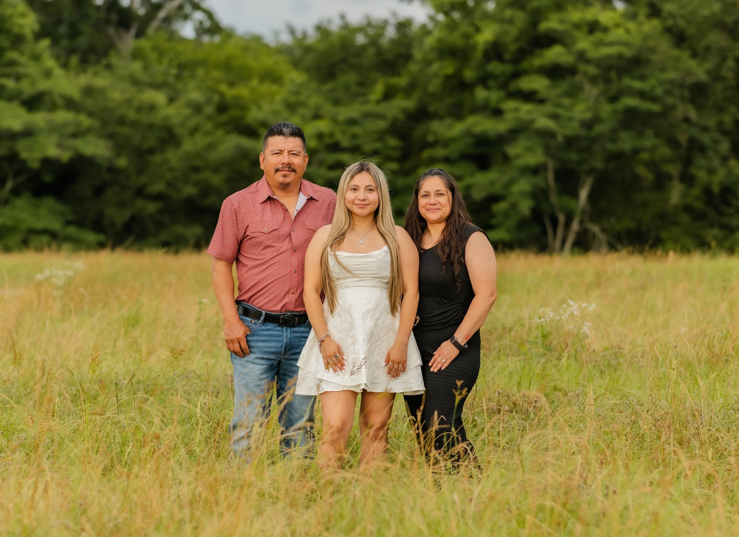 A family of three posing outdoors in a grassy field with green trees in the background.