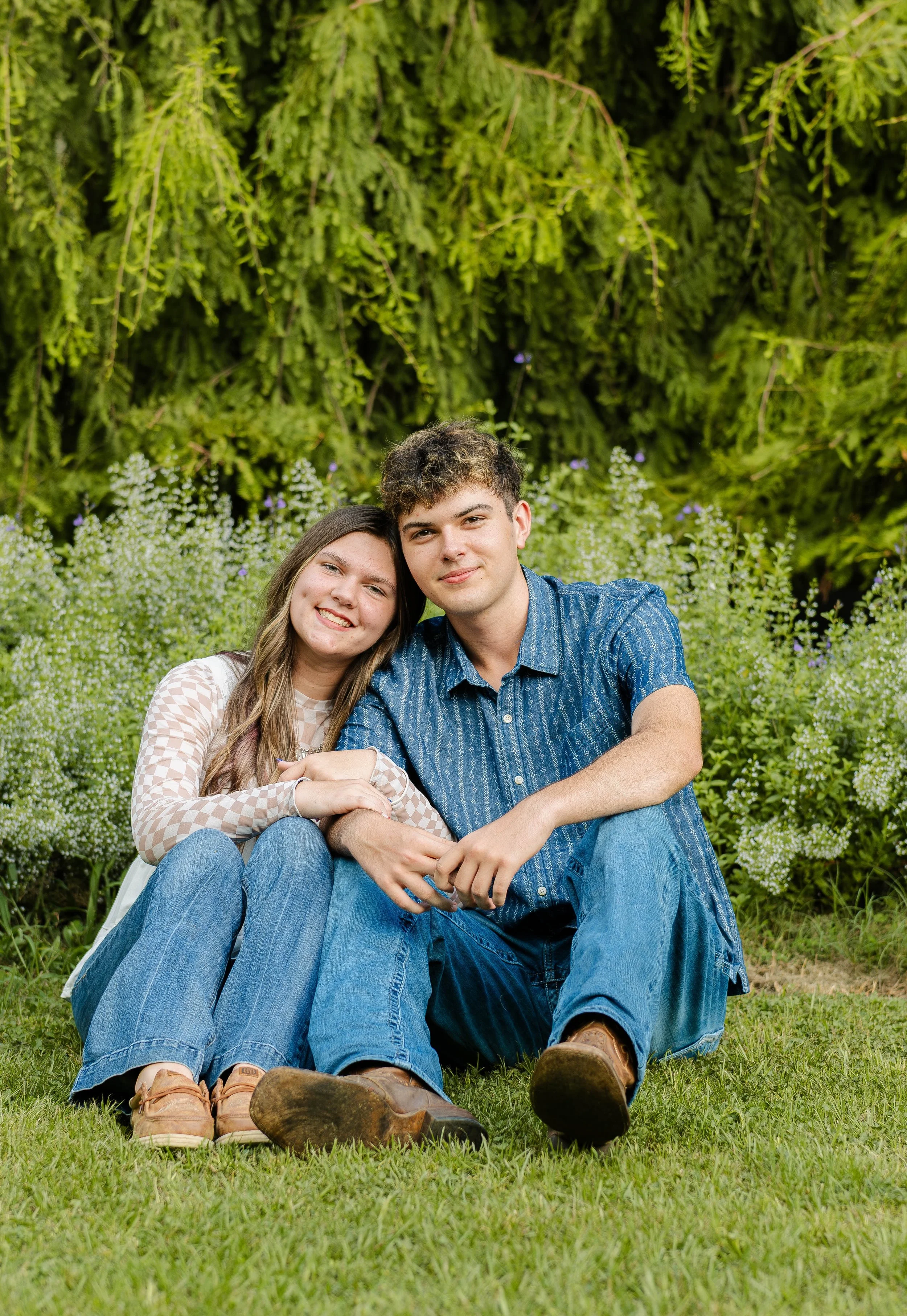 A young couple sitting on the grass in a garden with greenery and flowering bushes, smiling at the camera.