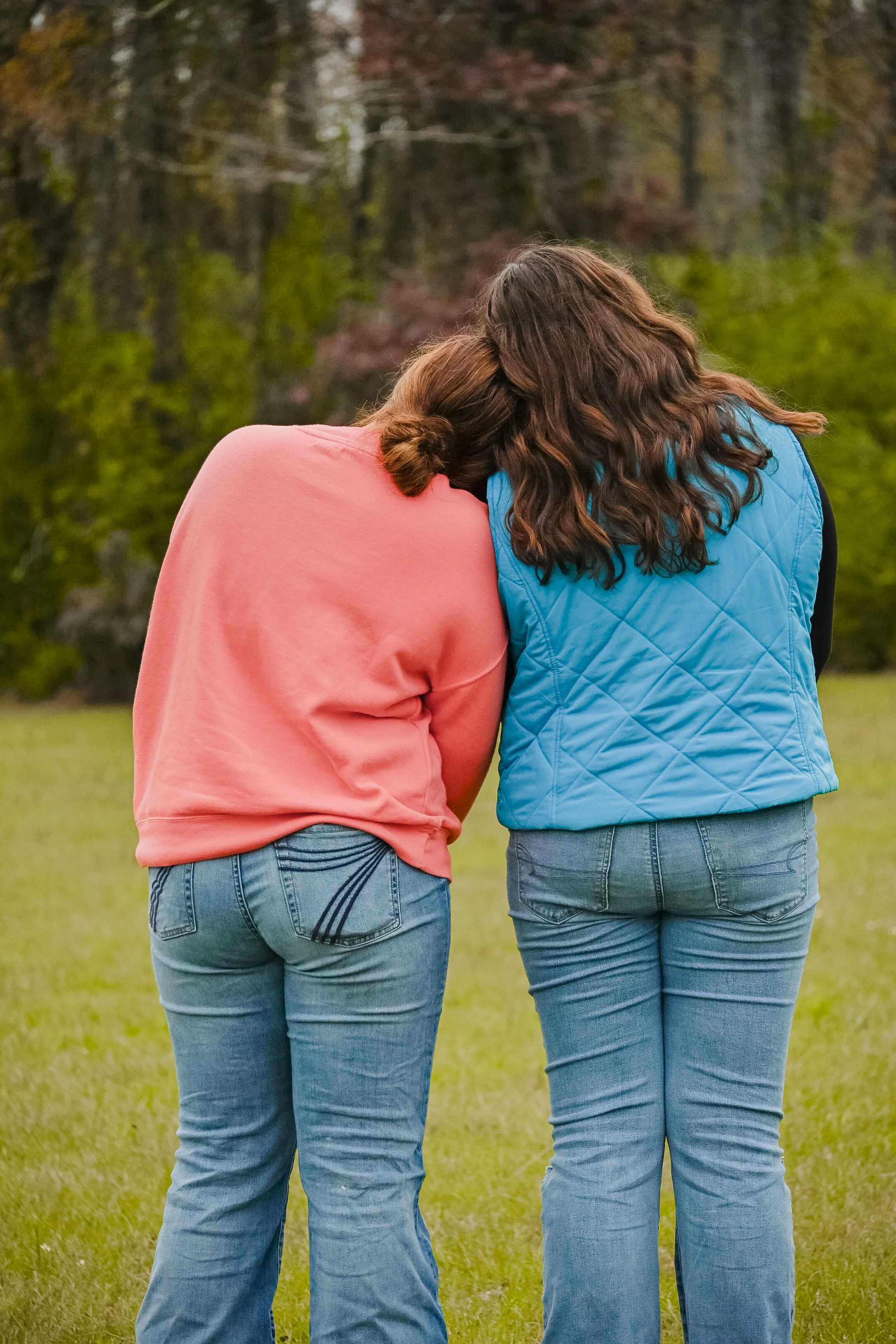 Two women standing in a grassy field with trees in the background, leaning their heads on each other's shoulders, wearing casual clothing.