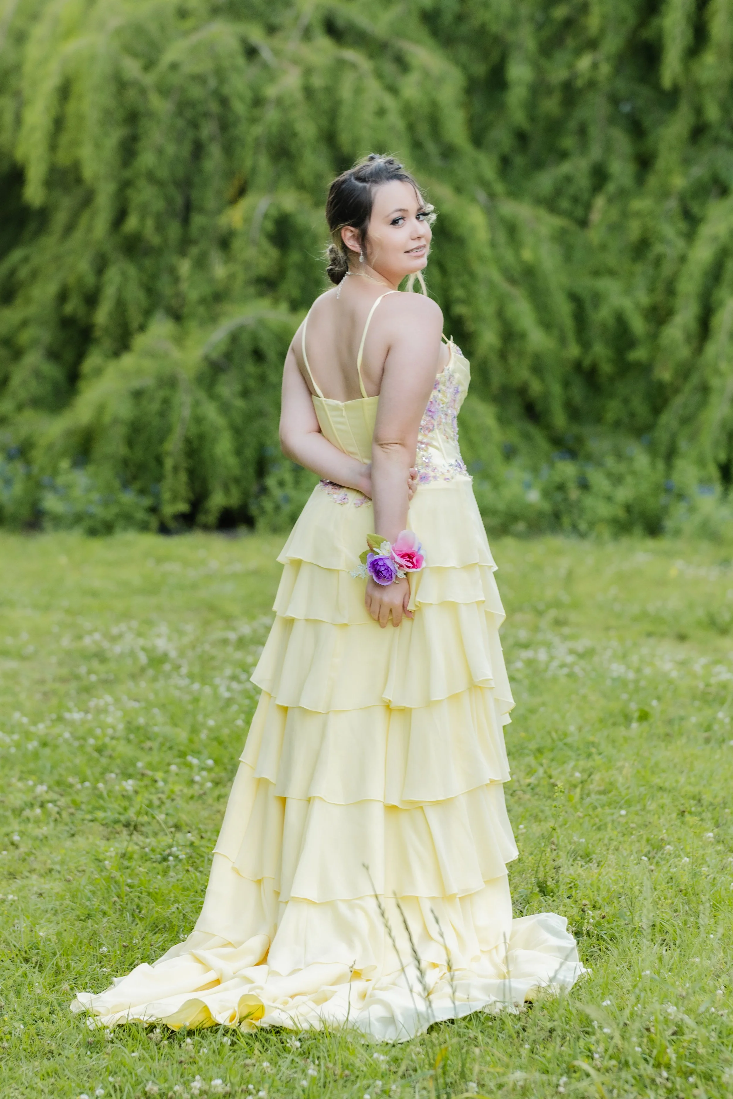A woman in a yellow dress standing outdoors on grass with trees in the background, holding a bouquet of flowers, and looking over her shoulder at the camera.