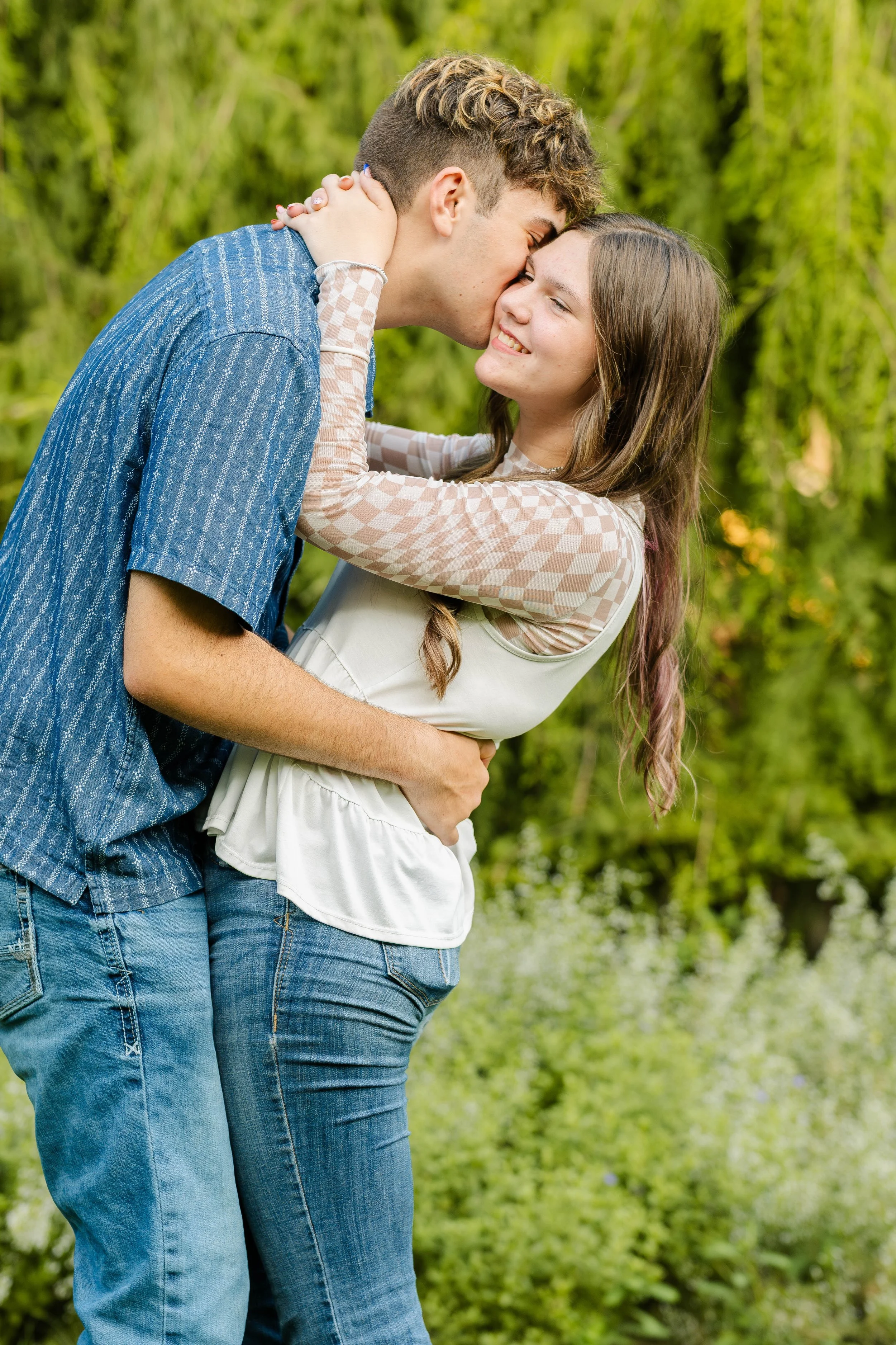 A young couple sharing a kiss outdoors, with the man kissing the woman's forehead, both smiling and surrounded by greenery.