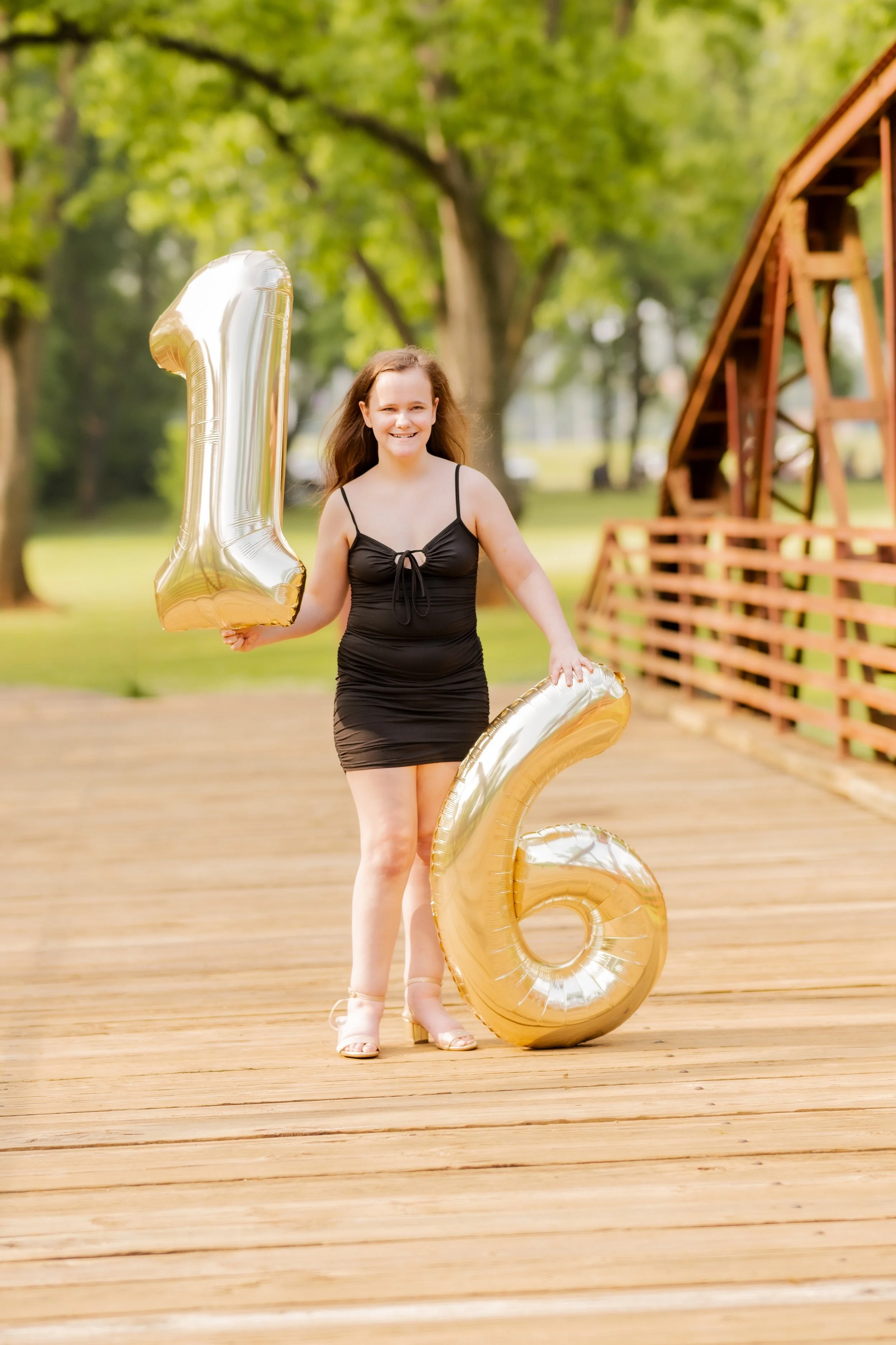 A young woman with brown hair in a black dress holding gold balloons shaped as the numbers one and six outdoors on a wooden deck, with green trees and a wooden structure in the background.