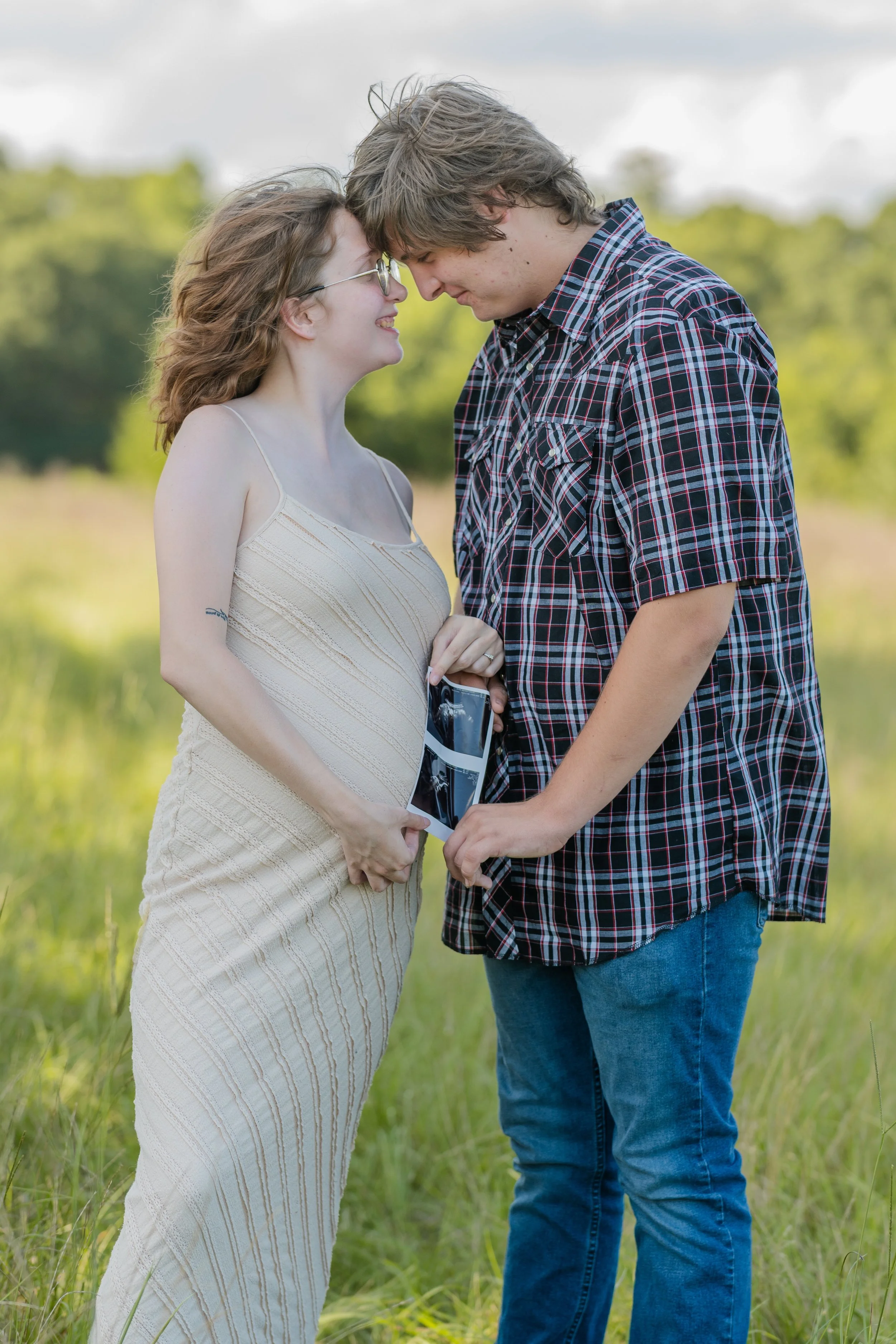 A young couple stands close, with foreheads touching, outdoors in a grassy field with trees in the background. The woman is pregnant, wearing a beige striped dress, and holding an ultrasound picture. The man is wearing a plaid shirt and jeans, and is holding her hand.