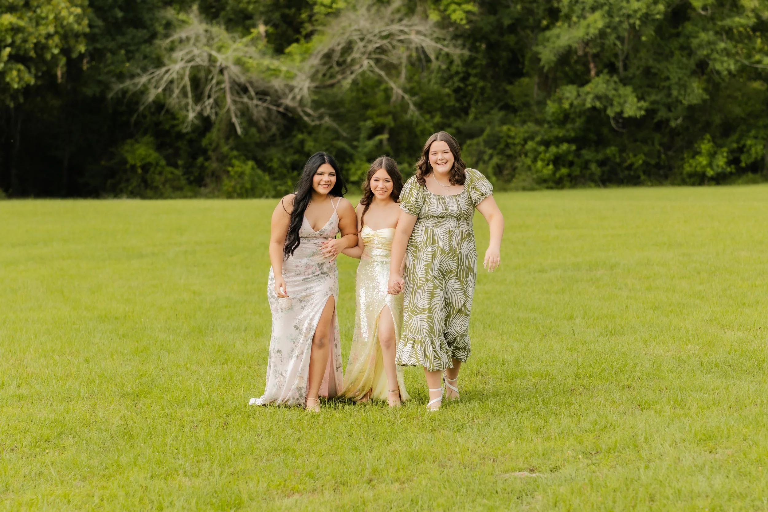 Three women walking hand in hand in a grassy field, smiling, with a background of green trees.