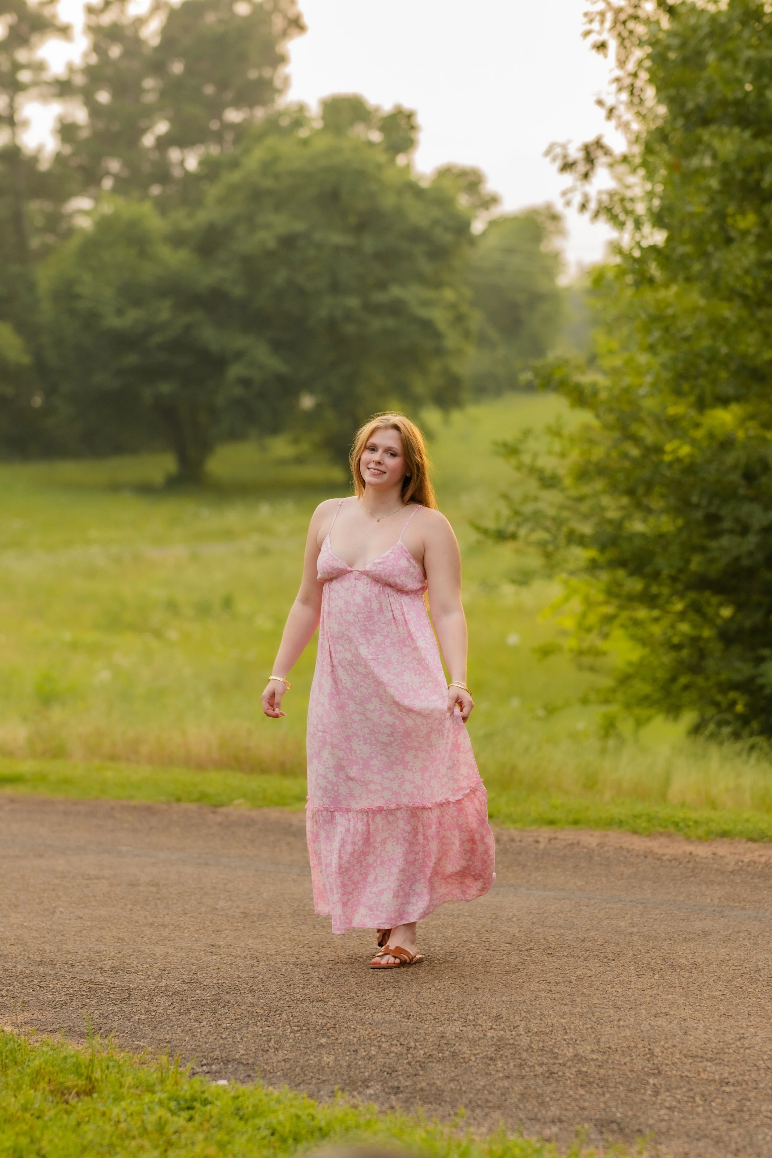A woman with long red hair in a pink dress walking on a gravel path in a lush green park during daylight.