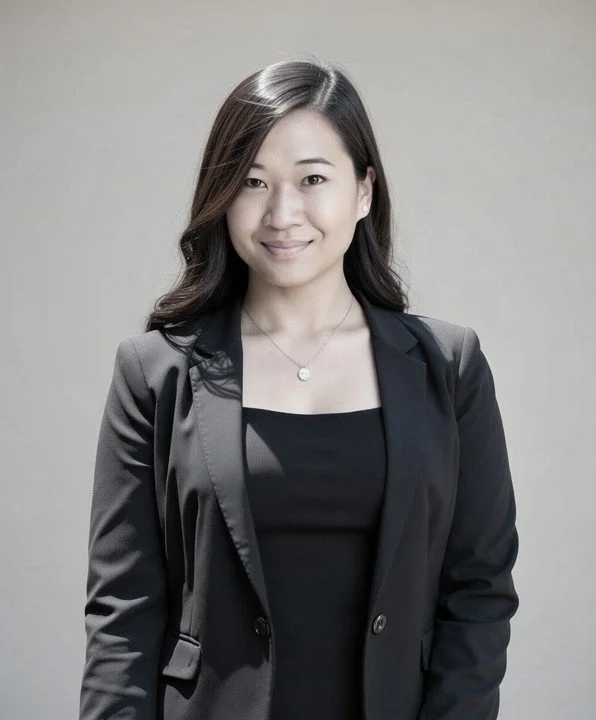 A professional Asian woman with shoulder-length dark hair, smiling, wearing a black blazer over a black top, and a silver necklace, standing in front of a neutral background.