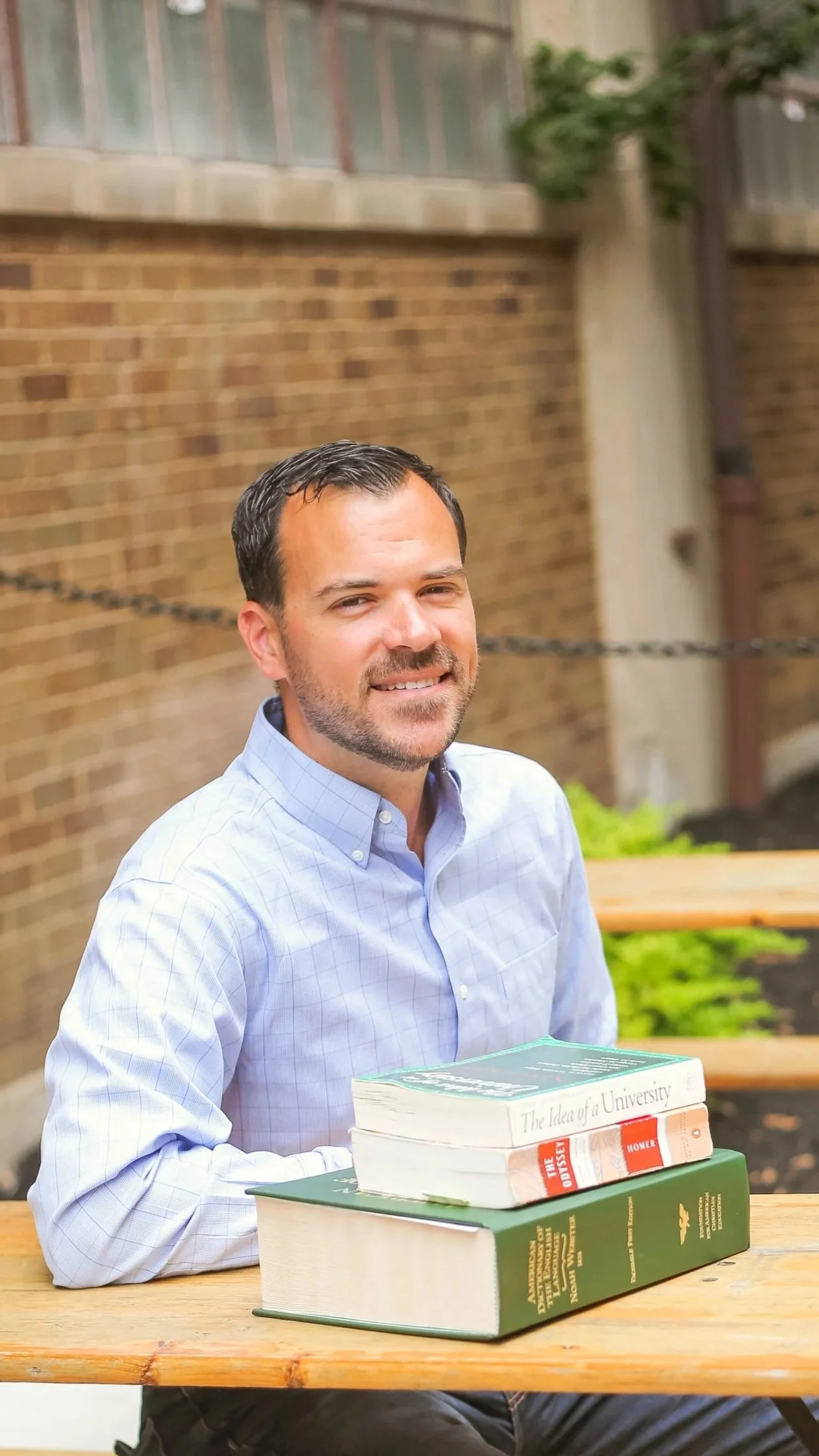 A man sitting at an outdoor wooden table with several books stacked in front of him, including titles like 'The Odyssey' and 'The Idea of a University,' in front of a brick wall and greenery.