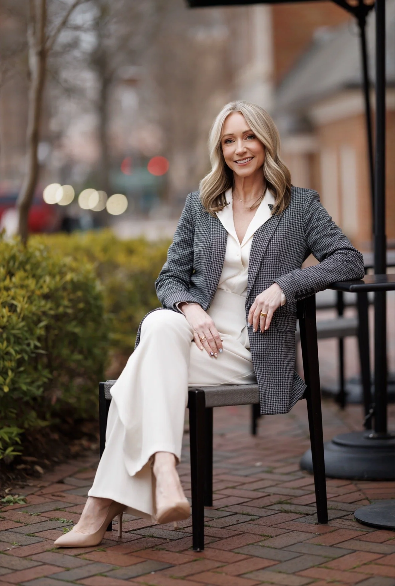A woman with blonde hair smiling and sitting outdoors on a chair near a table, dressed in a checkered blazer, white blouse, white wide-leg pants, and nude heels, with a blurred background of trees and buildings.
