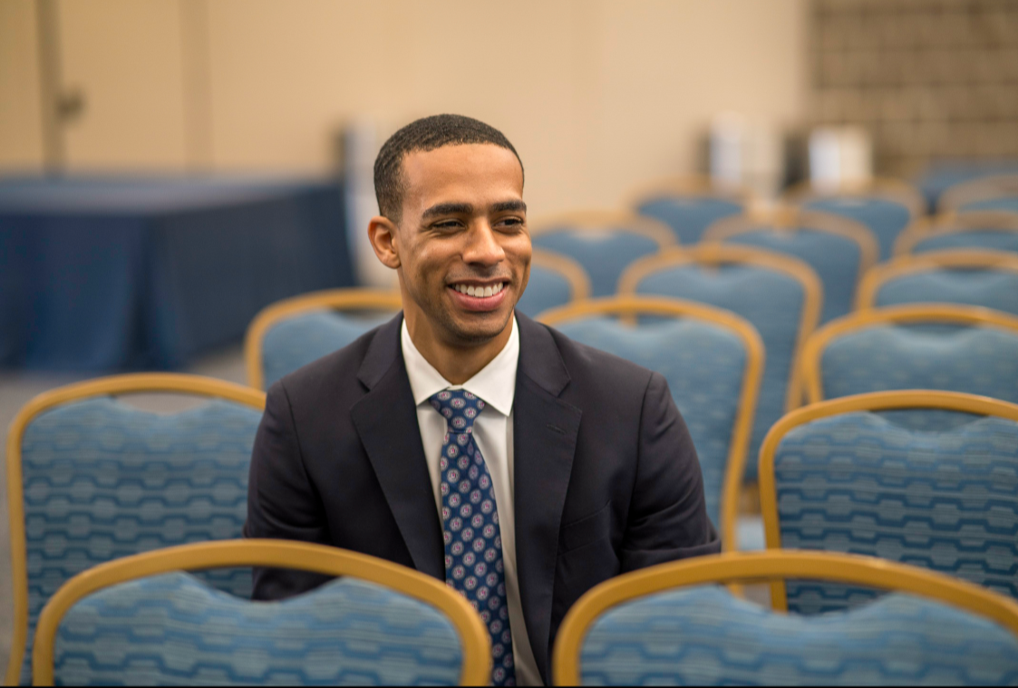 A smiling man in a suit sitting alone in an empty conference room with blue chairs.