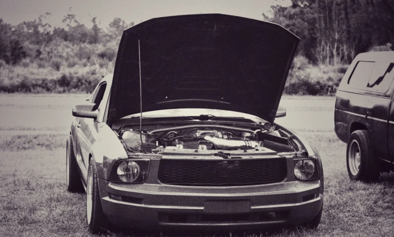 A black and white image of a vintage car with its hood open, parked on grass, with another vehicle parked nearby.