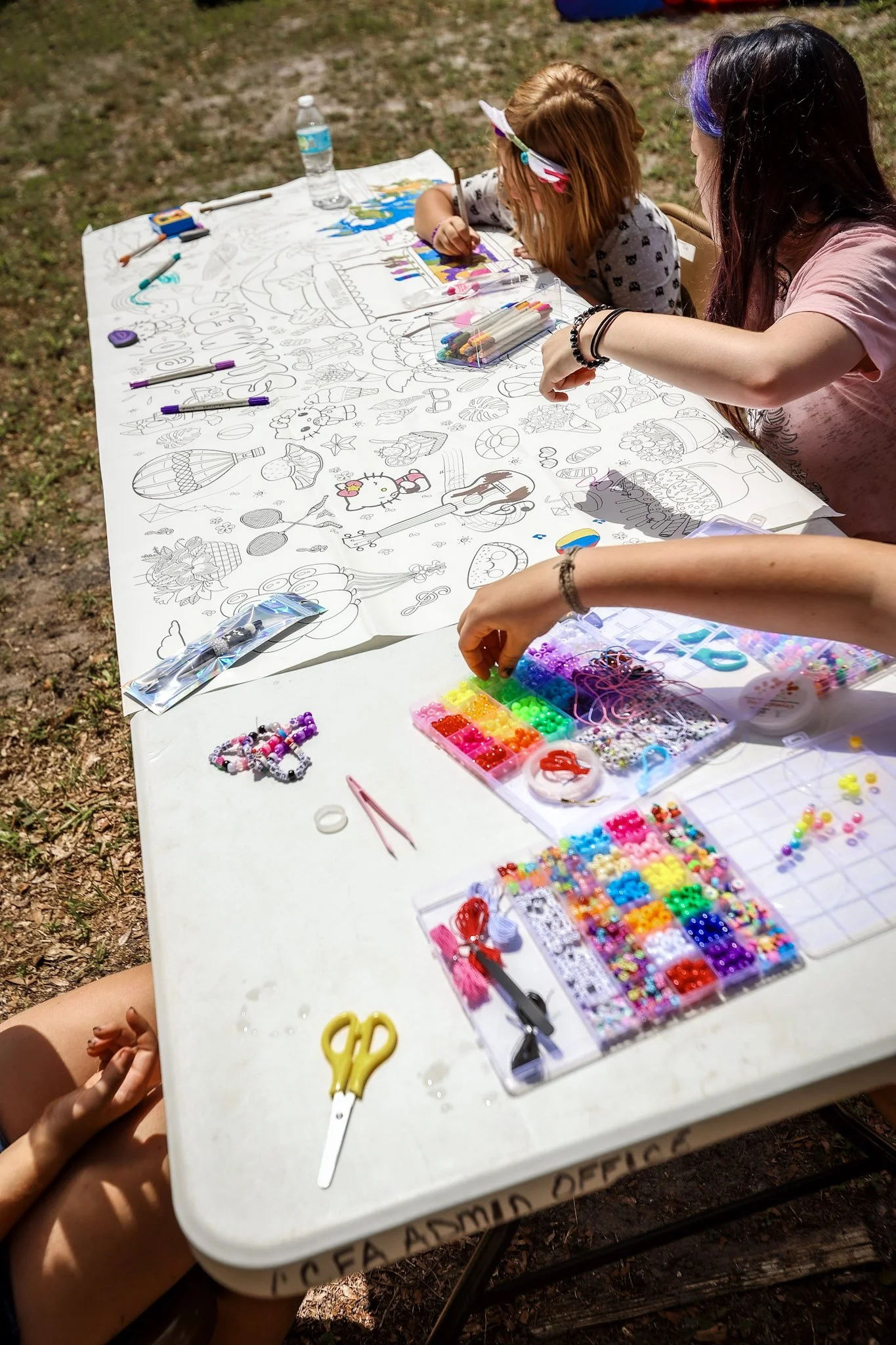 Children drawing on a large coloring poster with markers and beads, outside on a white table with various art supplies.