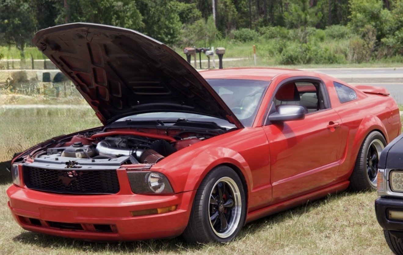 Red Ford Mustang with open hood parked on grass near road, surrounded by greenery.