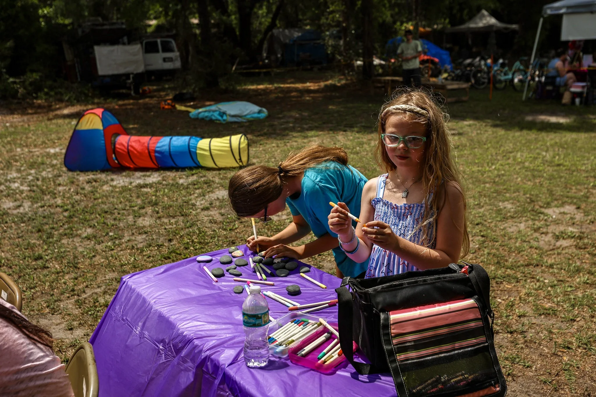 Children sitting at a purple-covered table outdoors with rocks, markers, and other craft supplies, surrounded by a camping area with tents, trees, and camping gear.