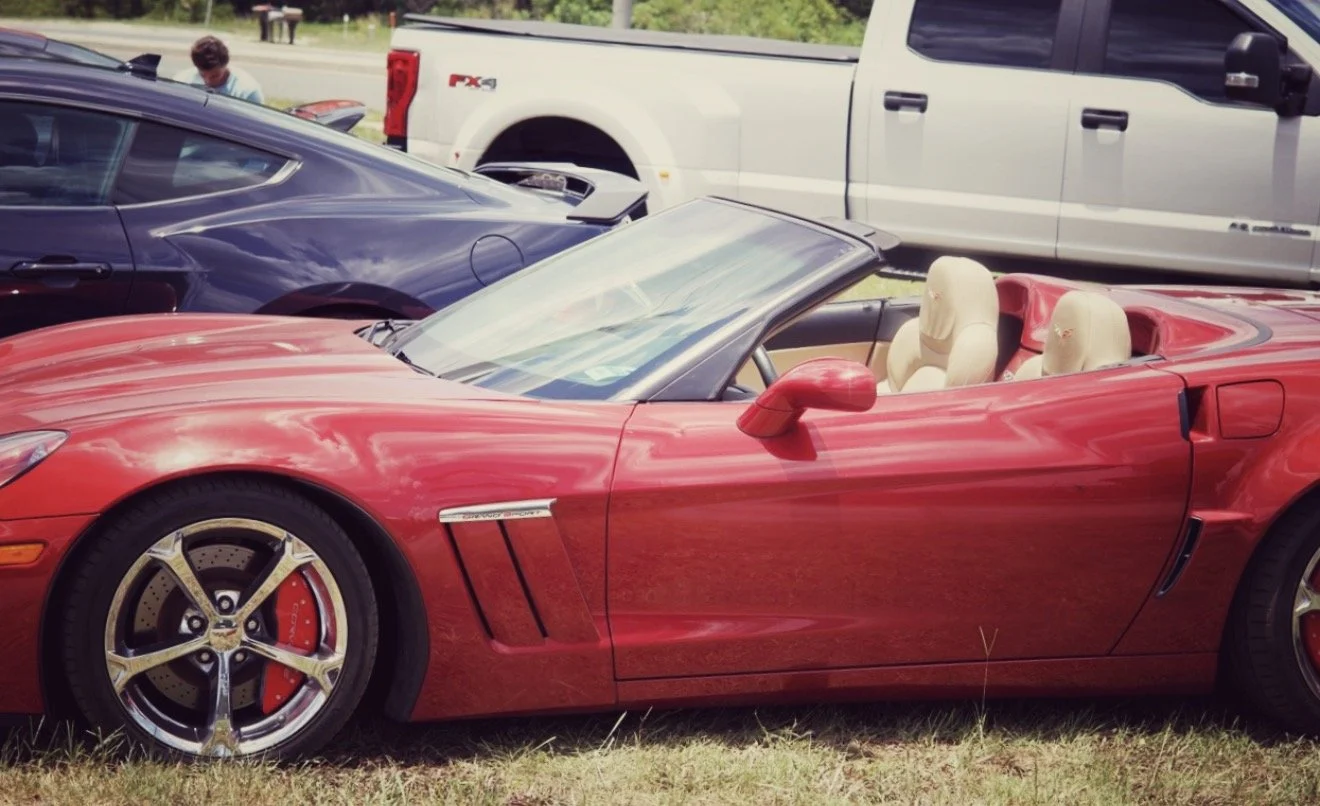 A red convertible sports car parked on grass, with a beige interior and two seats. There are other cars and a person in the background.