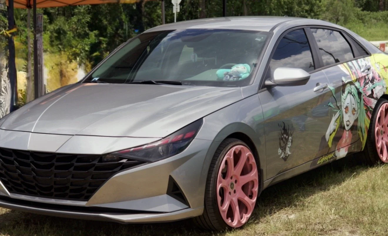 A silver car with pink rims parked on grass, decorated with anime-style character decals on the side and a plush toy on the dashboard.
