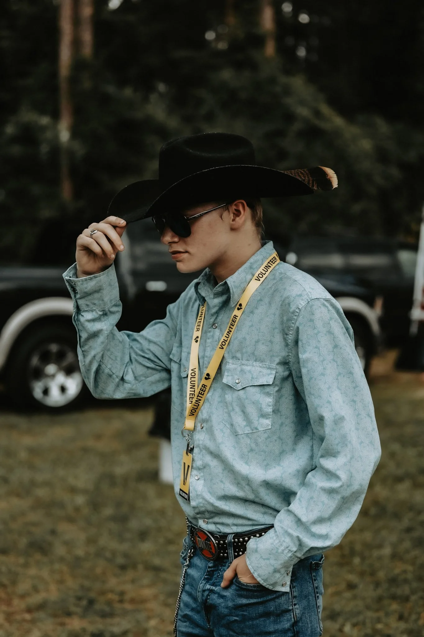 A man wearing a light blue western shirt with a patterned design, a black cowboy hat, sunglasses, a yellow volunteer lanyard, a large belt buckle, and jeans. He is outdoors with trees and vehicles in the background, tipping his hat.
