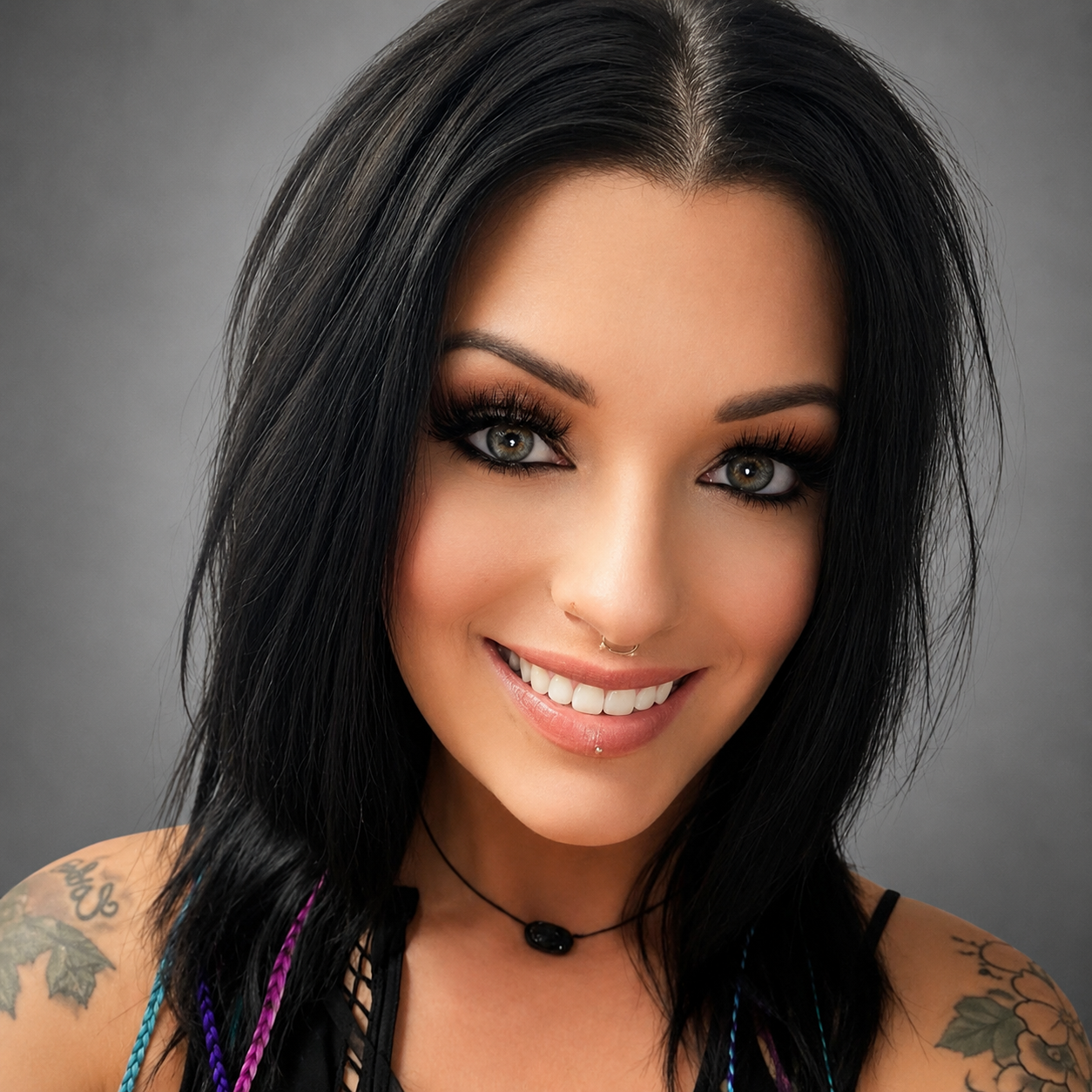 Close-up of smiling woman with black hair, gray eyes, tattoos on shoulders, and septum piercing, against gray background.
