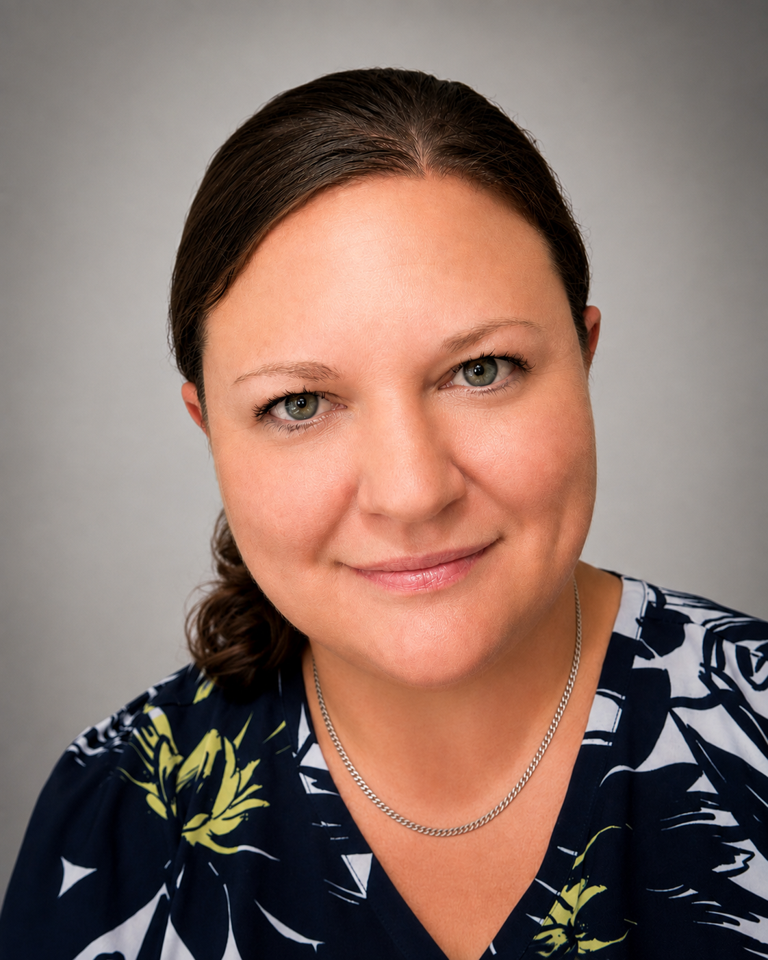 A close-up portrait of a woman with dark brown hair, blue eyes, and a subtle smile, wearing a navy blue top with yellow and white floral patterns and a silver necklace, against a plain gray background.