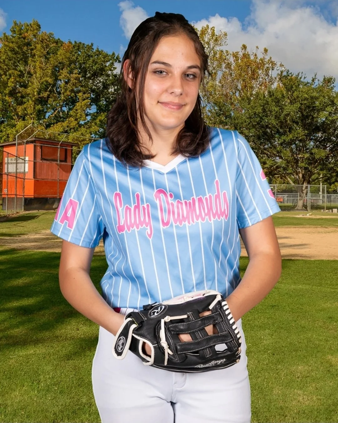 Teen girl wearing a light blue softball jersey with pink lettering that says 'Lady Diamonds' and holding a black softball glove, standing on a softball field with trees and a clear sky in the background.