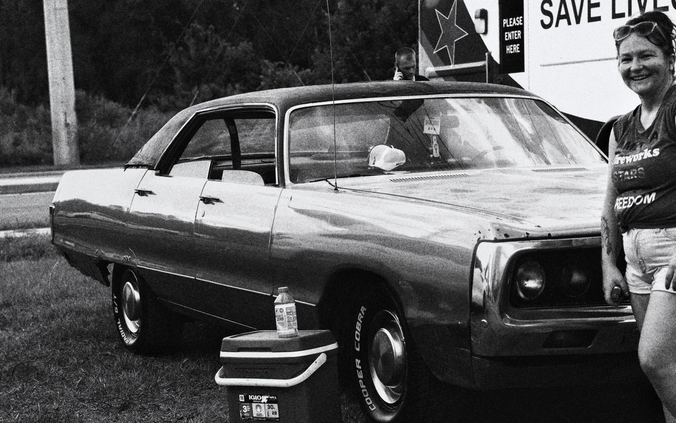 A vintage car parked on grass, with a woman smiling and standing to the right. There is a cooler, a water bottle, and a person talking on a cell phone in the background. A sign in the background reads 'Save Lives' and 'Please Enter Here.'