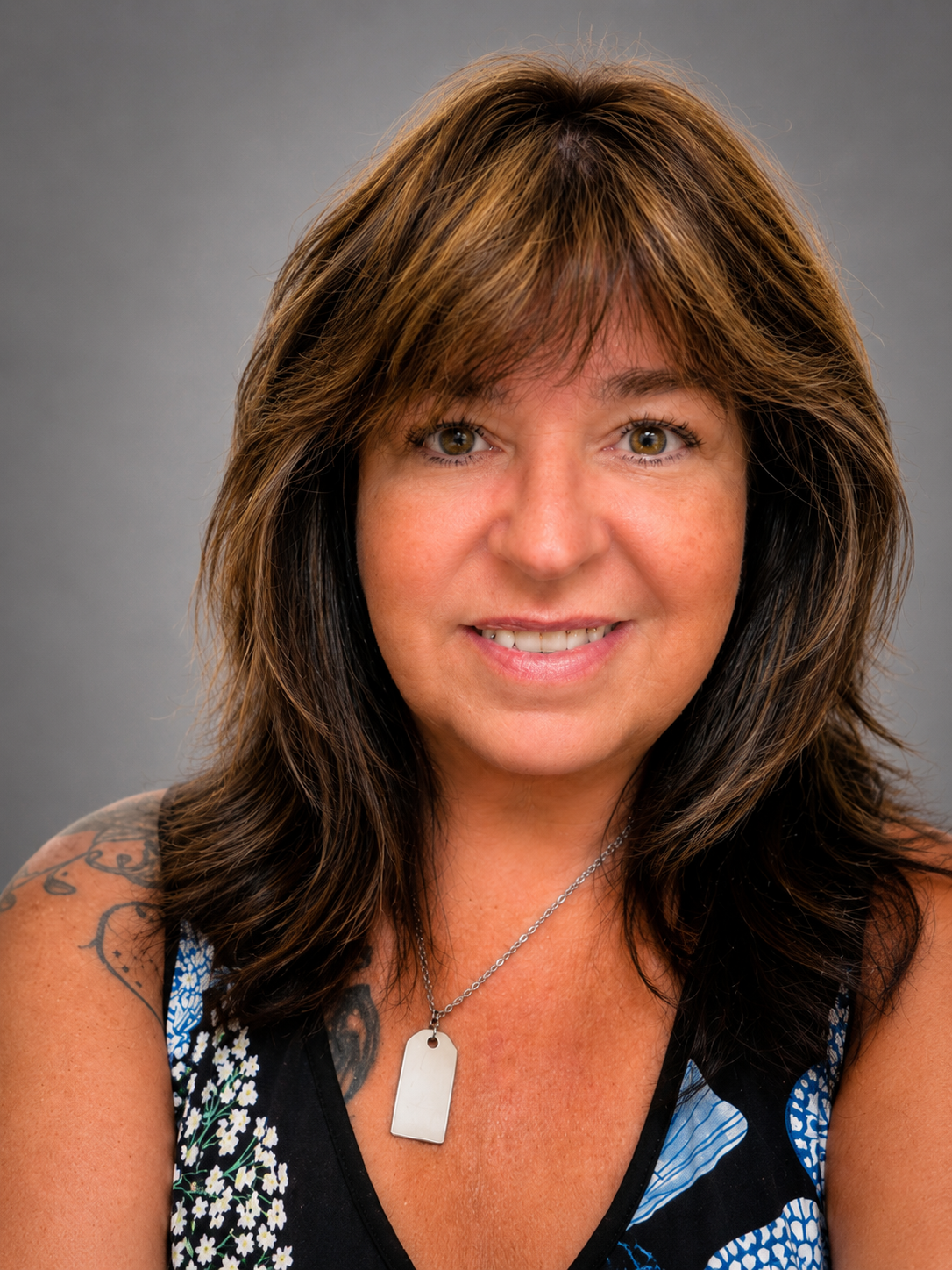 Close-up portrait of a smiling woman with shoulder-length brown hair and green eyes, wearing a black top with floral and blue patterns, and a silver necklace with a rectangular pendant, against a gray background.