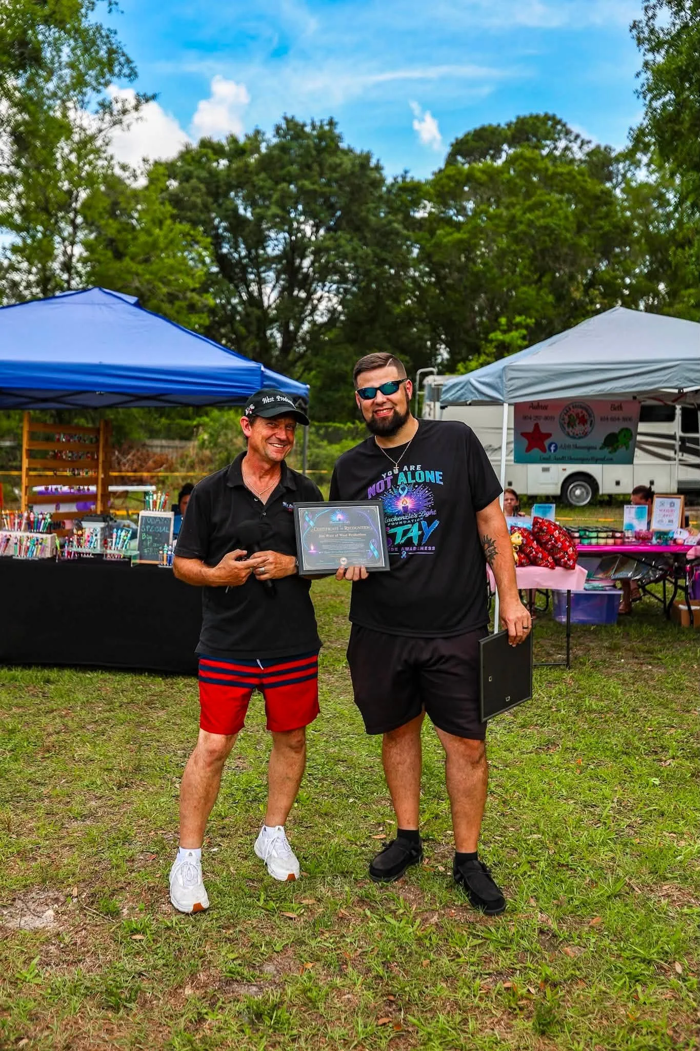 Two men standing outdoors at a park or festival, smiling and holding a certificate. They are wearing sunglasses and casual clothing. Behind them are tents and a table with items on display, and trees under a blue sky.