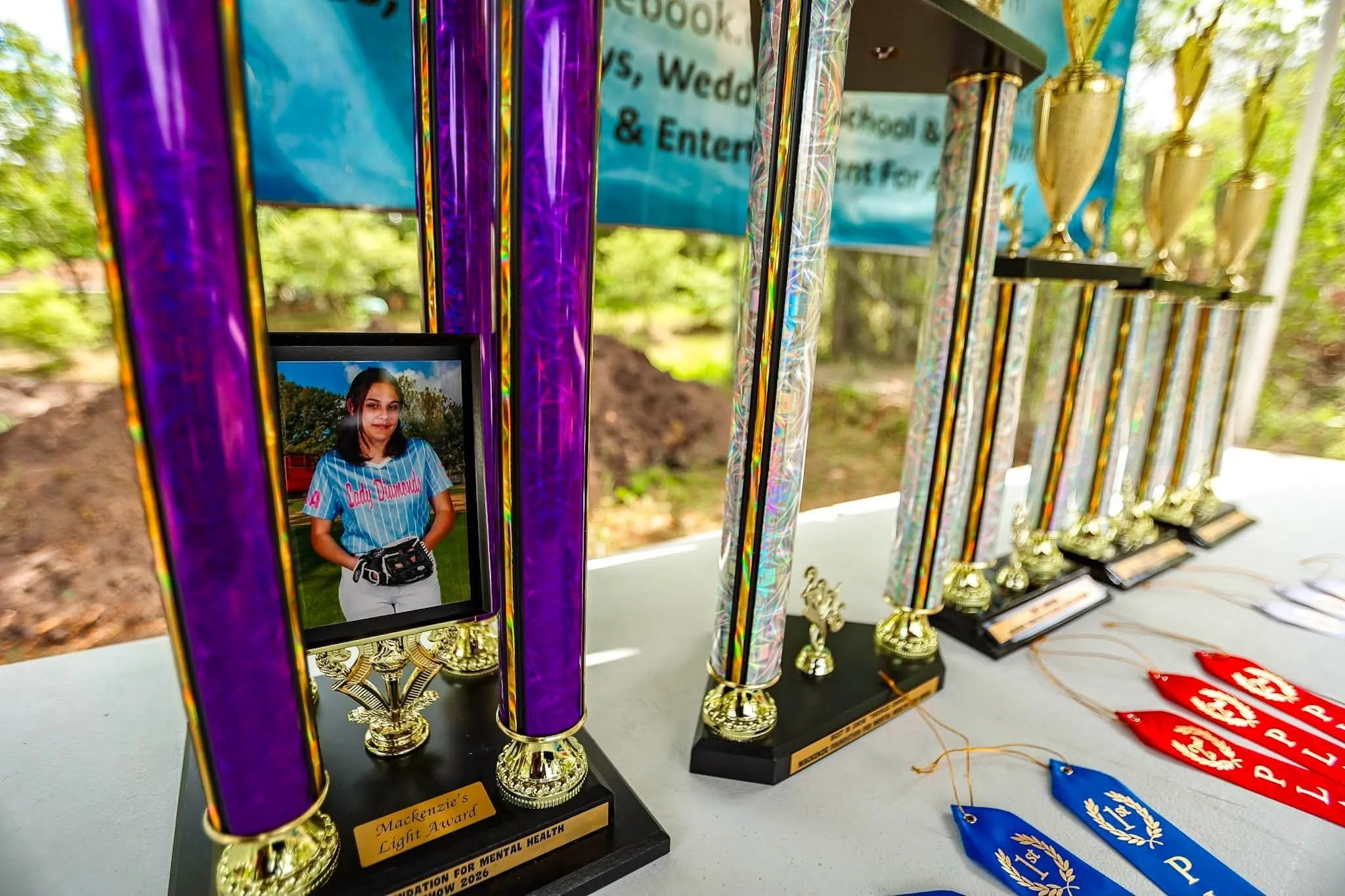 Photograph of various awards and trophies displayed on a table outdoors. The trophies are tall, colorful, and shiny, with some featuring gold cups and others with plaques. One of the awards has a picture of a young girl in a baseball uniform. Blue, r