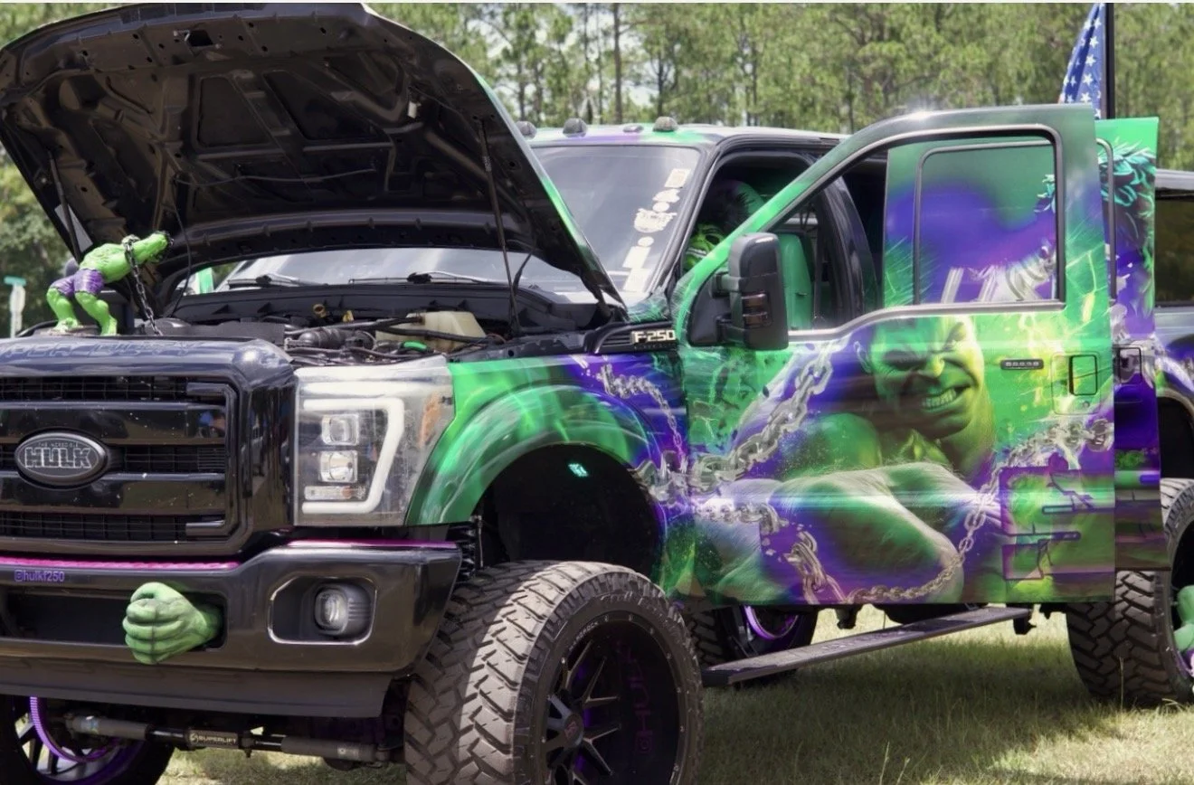 A decorated black Ford truck with a vibrant Hulk-themed paint job, featuring Hulk smashing through chains and smiling. The hood is open with green Hulk toys hanging from it, and the truck has large tires, purple accents, and an American flag in the b