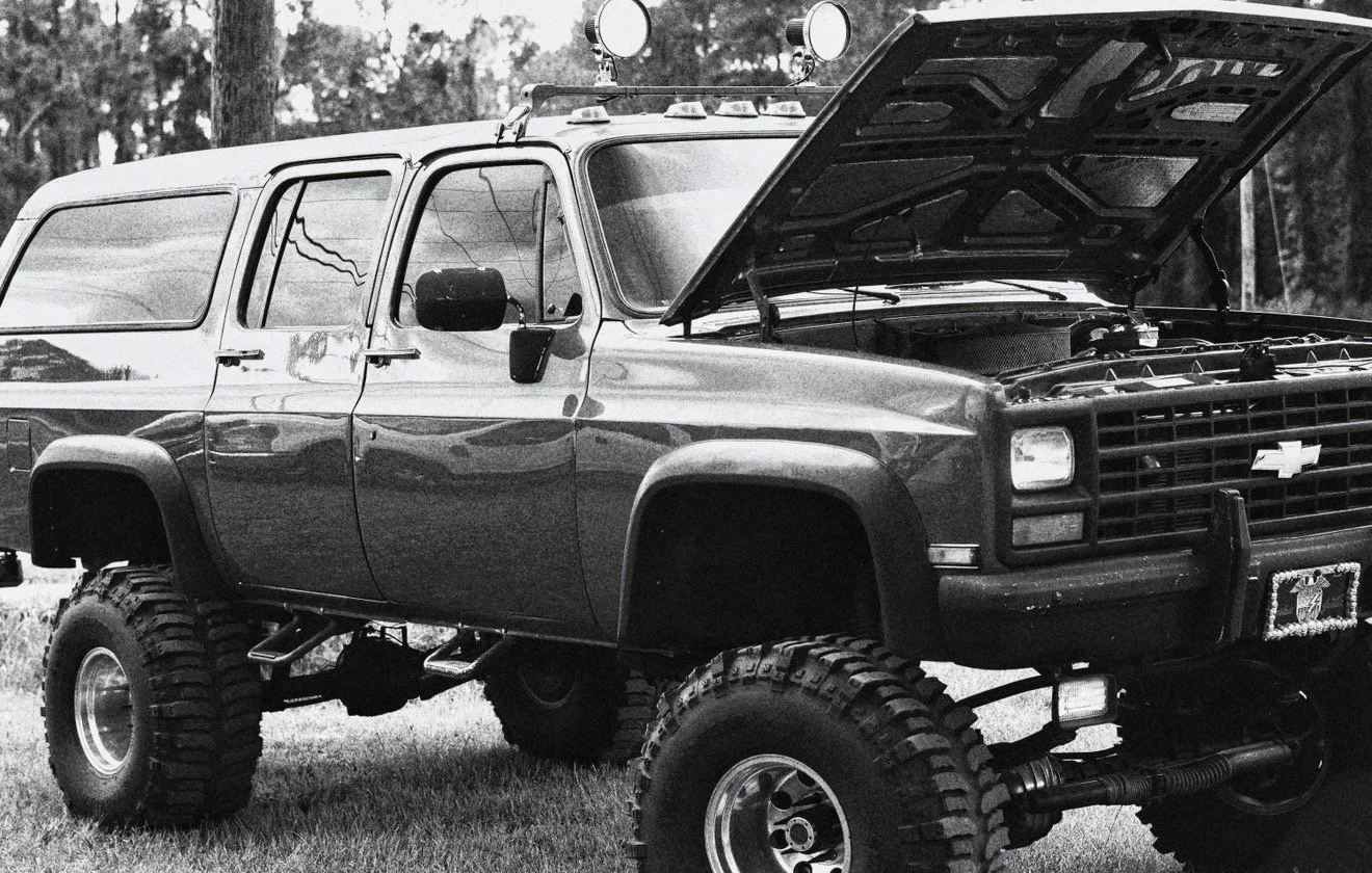 A black and white photo of a lifted Chevrolet SUV with its hood open, showing large off-road tires, a roof light bar, and a custom grille.