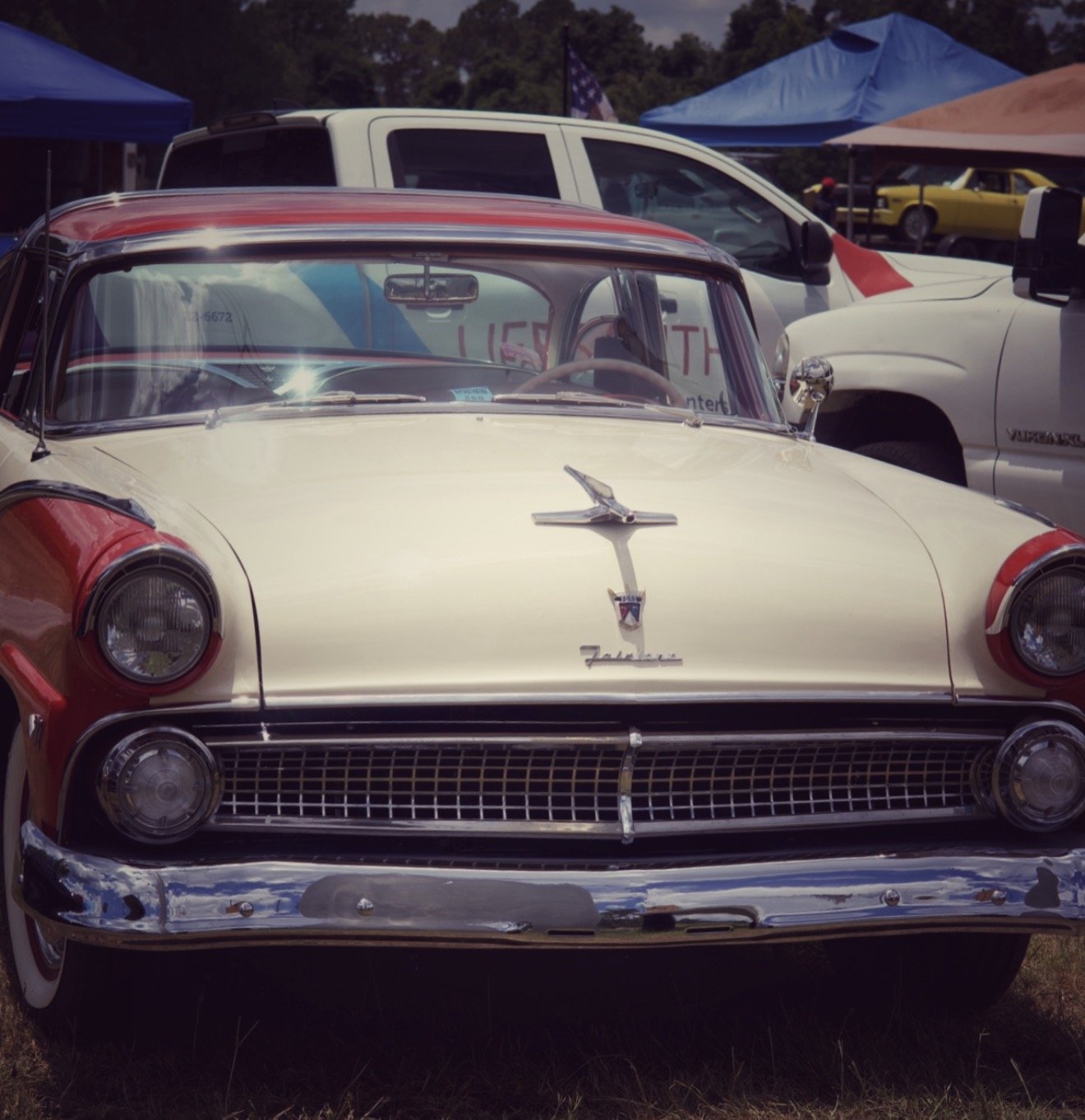 A vintage white and red Ford Thunderbird car with a chrome grille and classic design, parked at an outdoor car show with tents and other vehicles in the background.