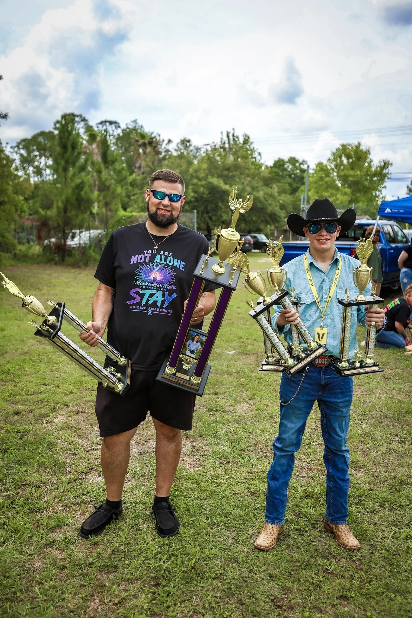 Two men standing outdoors on grass, holding trophies and smiling. The man on the left wears a black T-shirt, black shorts, black shoes, and sunglasses. The man on the right wears a cowboy hat, sunglasses, a patterned shirt, jeans, and cowboy boots. T