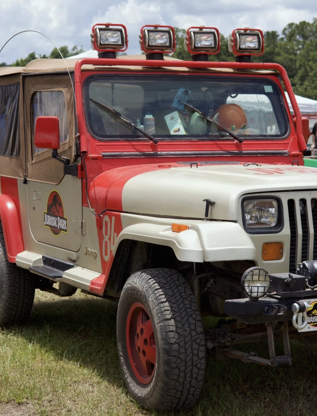 A Jeep vehicle decorated with Jurassic Park logos, labeled as '88, Jeep, and Jurassic Park,' equipped with four red lights on the roof, parked on grass, with tents and trees in the background.