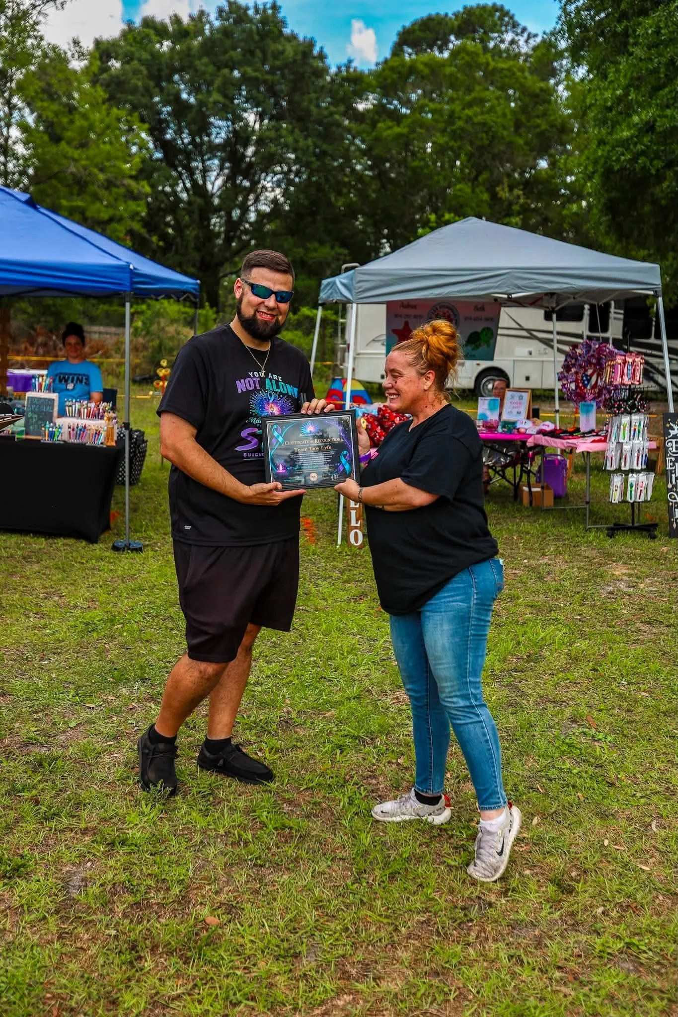 A man and woman smiling and holding a framed certificate at an outdoor event with vendor tents in the background.