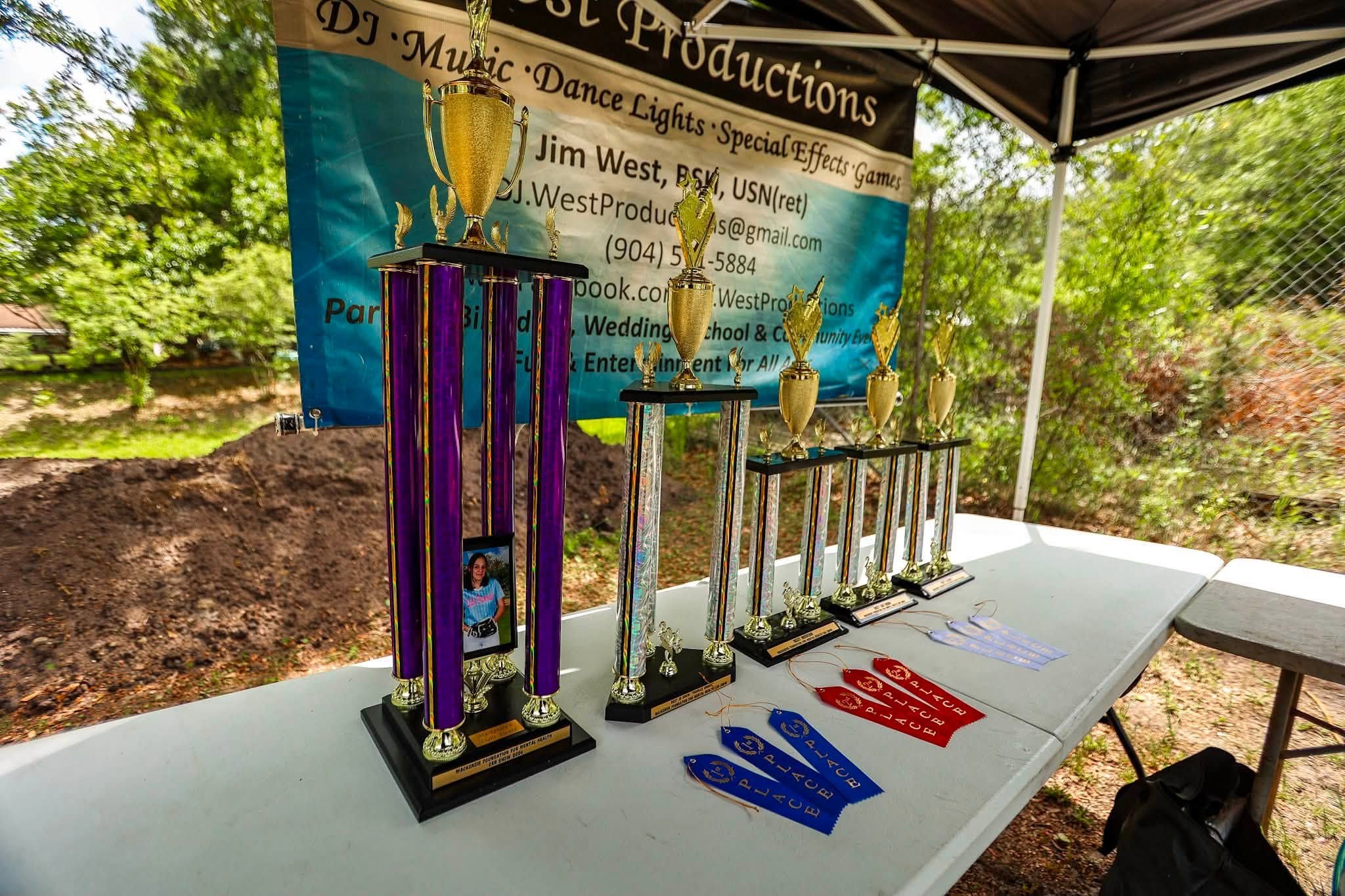 Several trophies and medals displayed on a white table at an outdoor event, with a banner in the background promoting music, dance, effects games, and entertainment.