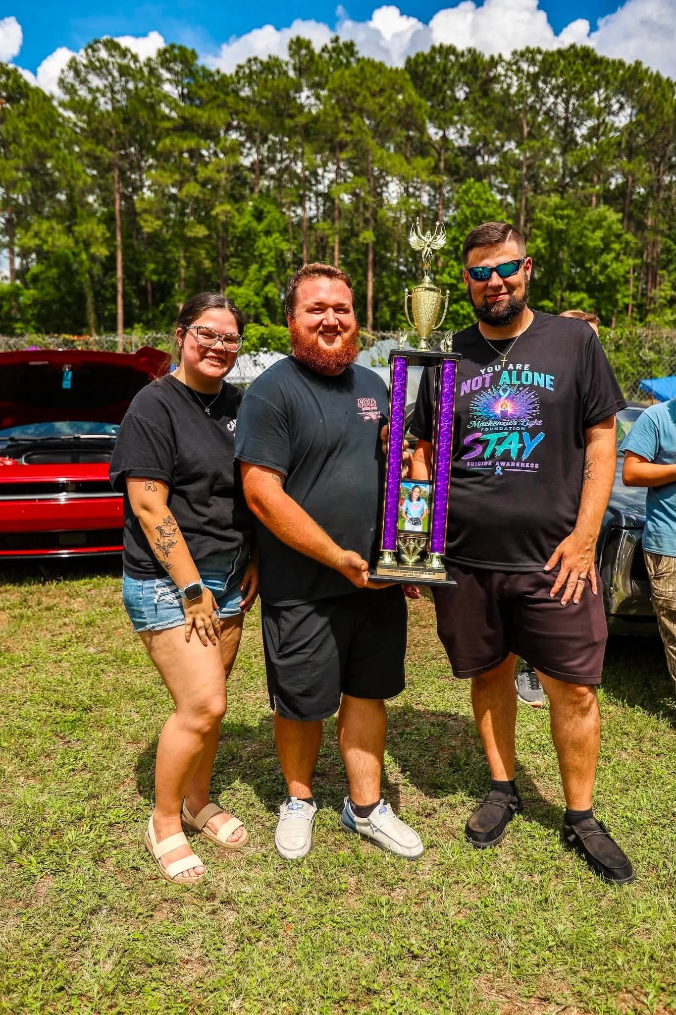 Three people standing outdoors on grass, holding a tall trophy, with a backdrop of trees and a sky with clouds. Two men and one woman smiling, with a red car partially visible behind them.