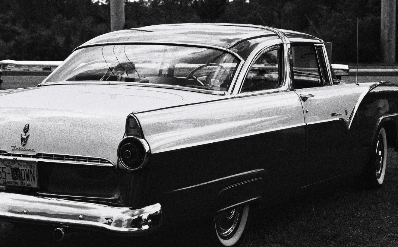 A vintage black and white car, likely from the 1950s, parked outdoors with trees in the background.