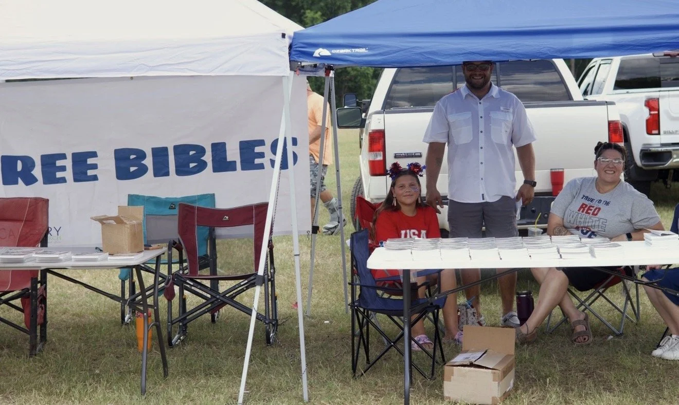 People sitting at a table outdoors at a Christian event. There is a banner reading "FREE BIBLES" behind them. Two women are seated, and one man is standing. One girl is sitting, all smiling.