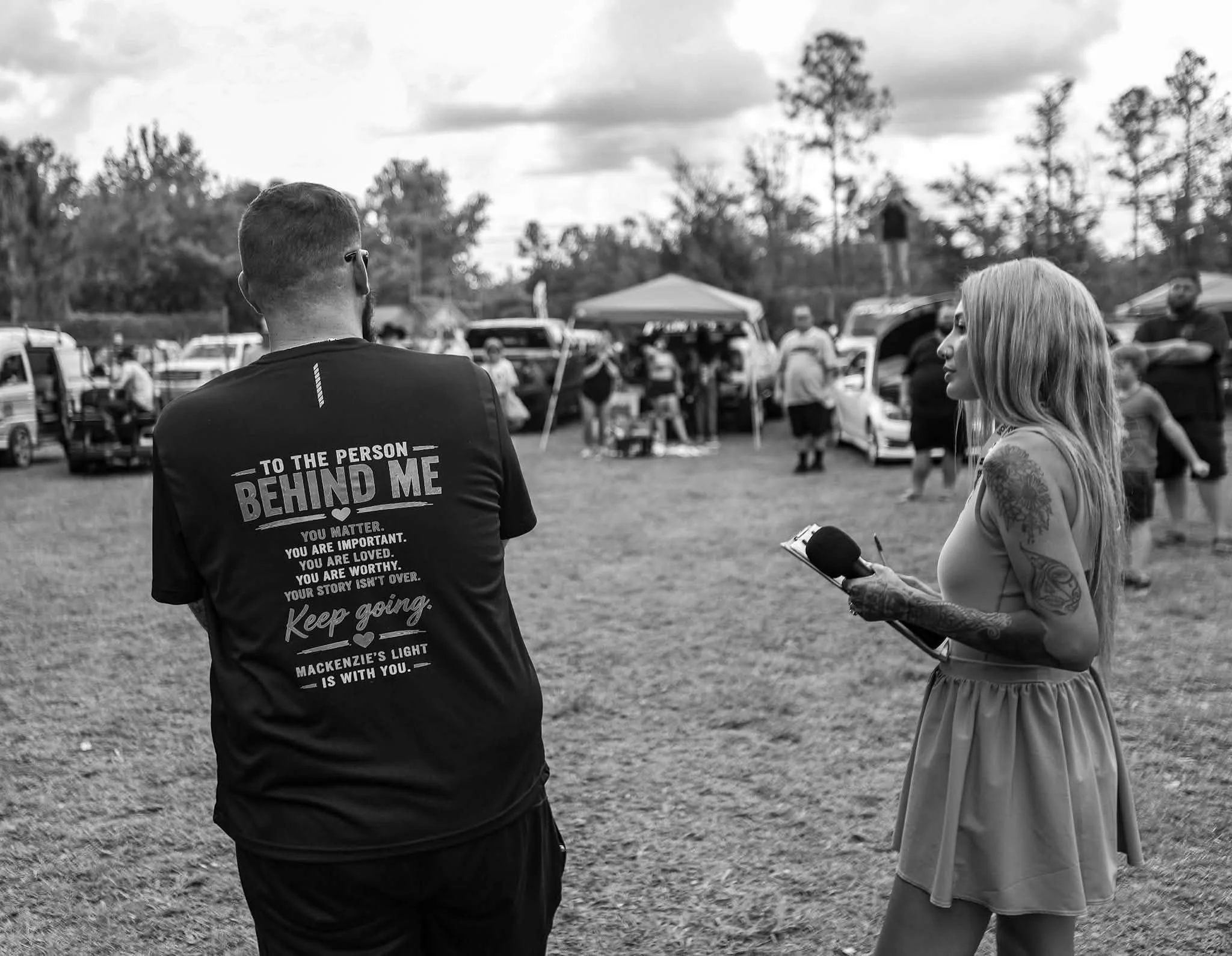 A black and white photo showing a young woman with long hair and tattoos on her arm holding a microphone, engaged in an interview at a crowded outdoor event. A man with his back to the camera wearing a T-shirt with motivational text is partially visible. Multiple cars and people are in the background with trees and a cloudy sky overhead.