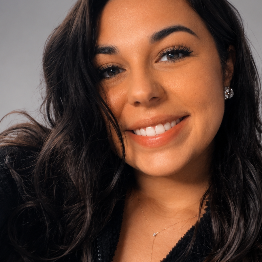 Close-up portrait of a woman with dark wavy hair, wearing makeup, diamond earrings, a gold necklace, and a black top, smiling at the camera.