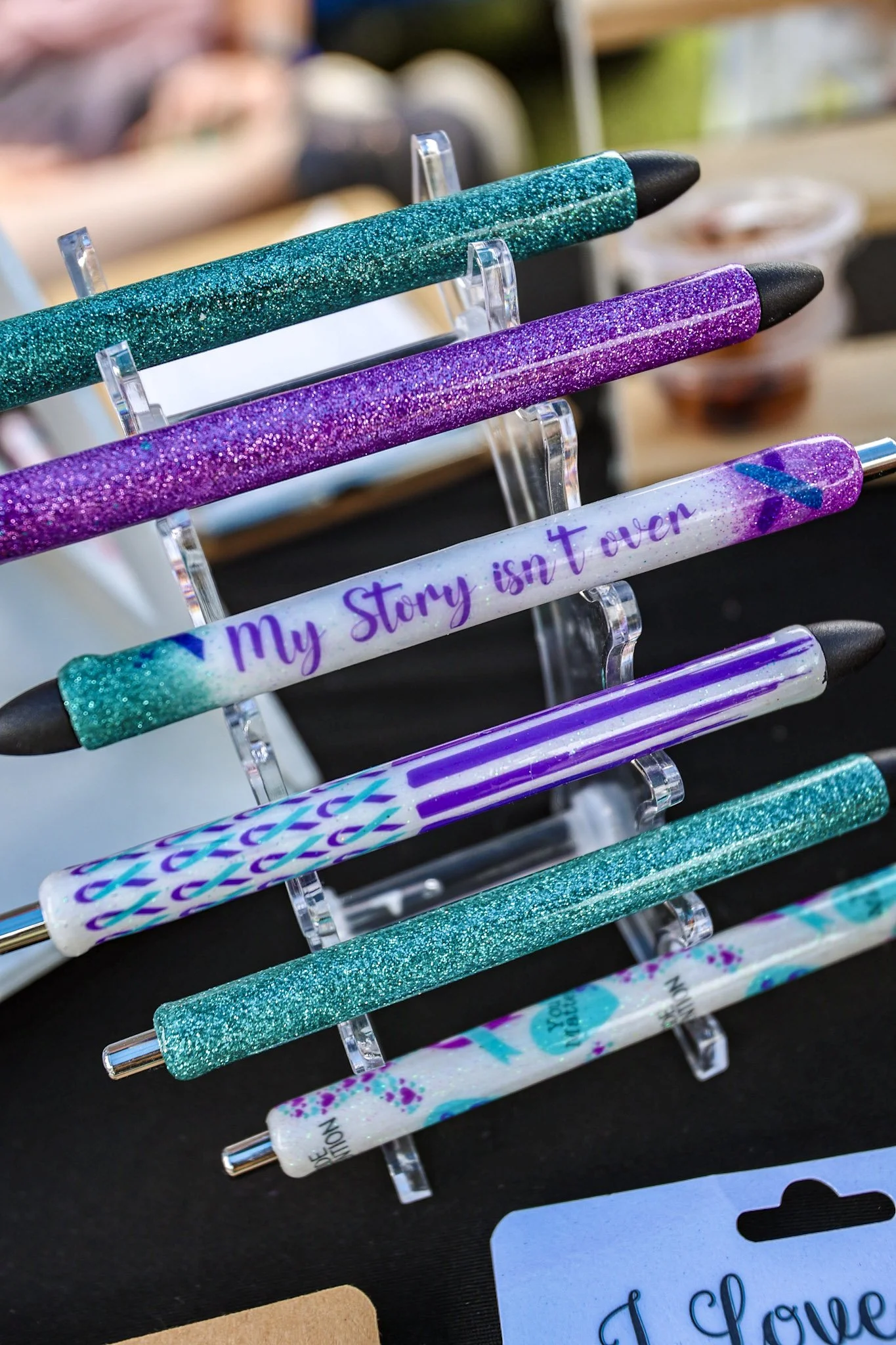 Close-up of sparkling pens with various colors and designs, displayed on a stand at a craft or stationery store.