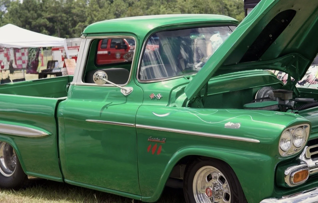 Green vintage pickup truck with open hood at an outdoor car show, tents and other vehicles in the background.