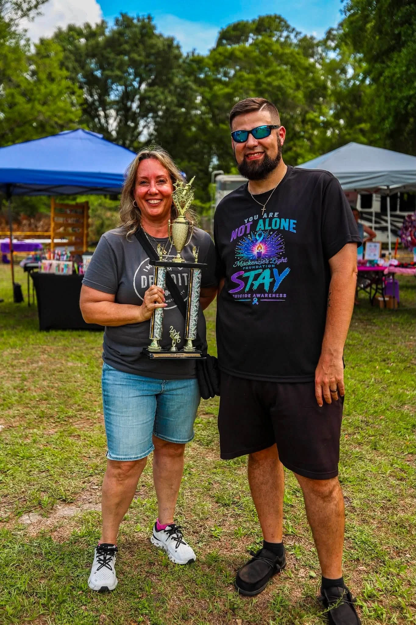 Two smiling people standing outdoors, holding a trophy. The woman on the left wears a gray t-shirt, denim shorts, and white sneakers with pink accents. The man on the right wears a black t-shirt with a Genesis Light Foundation message, black shorts, 