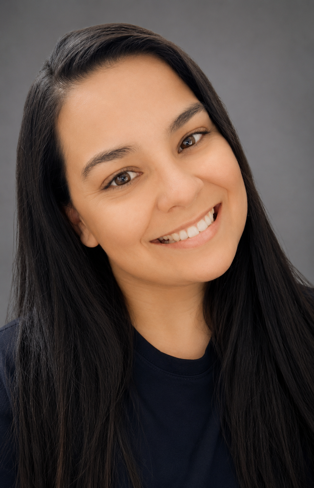 Close-up portrait of a young woman with long dark hair smiling against a plain gray background.