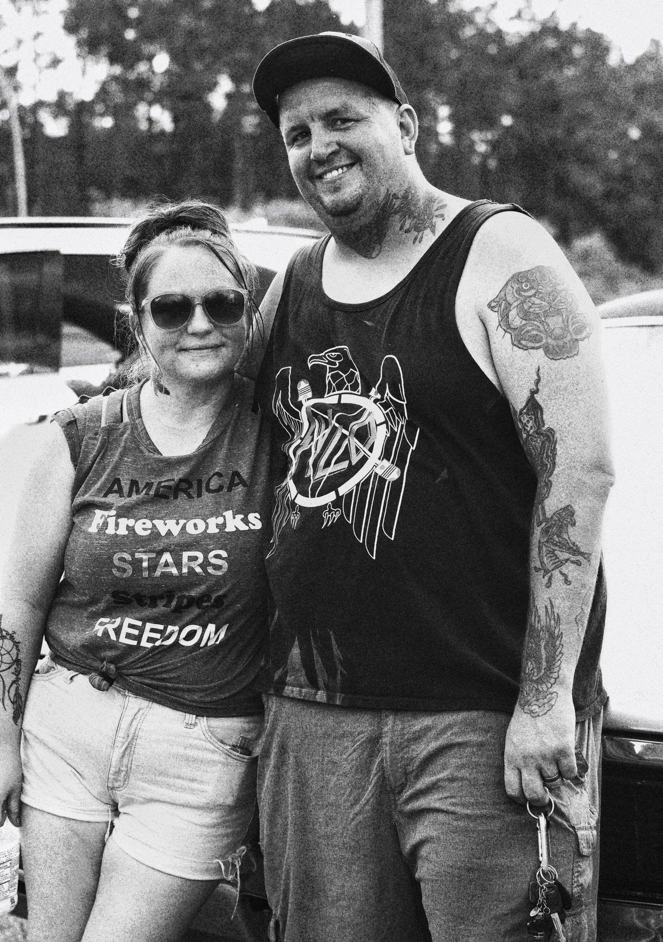 A smiling man and woman posing outdoors. The woman wears sunglasses, a t-shirt with patriotic words, and distressed shorts. The man wears a tank top with a graphic and has tattoos on his arms and neck.