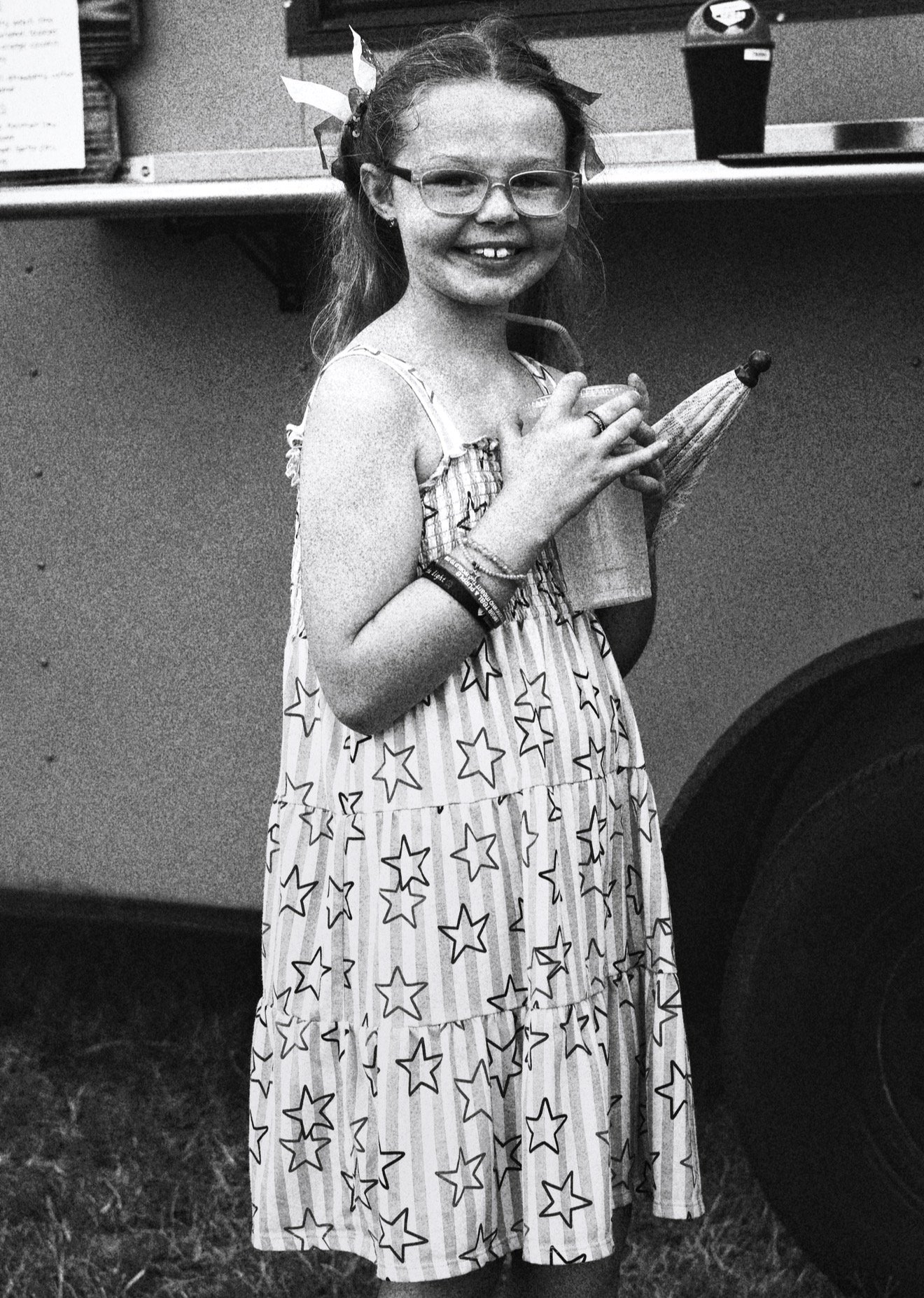 A young girl with glasses and a dress with star and stripe patterns, holding a small umbrella, smiling, standing outside near a car with a potted plant on the trunk in the background.